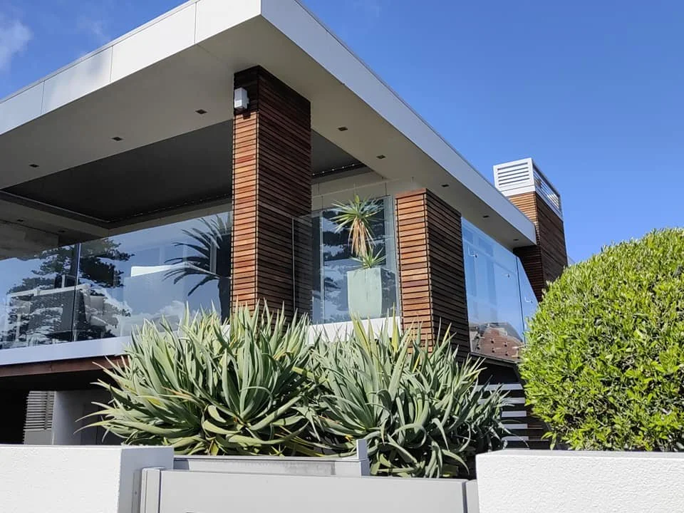 Modern house with large glass windows, wooden accents, and plants outside, including a potted plant and a green bush.