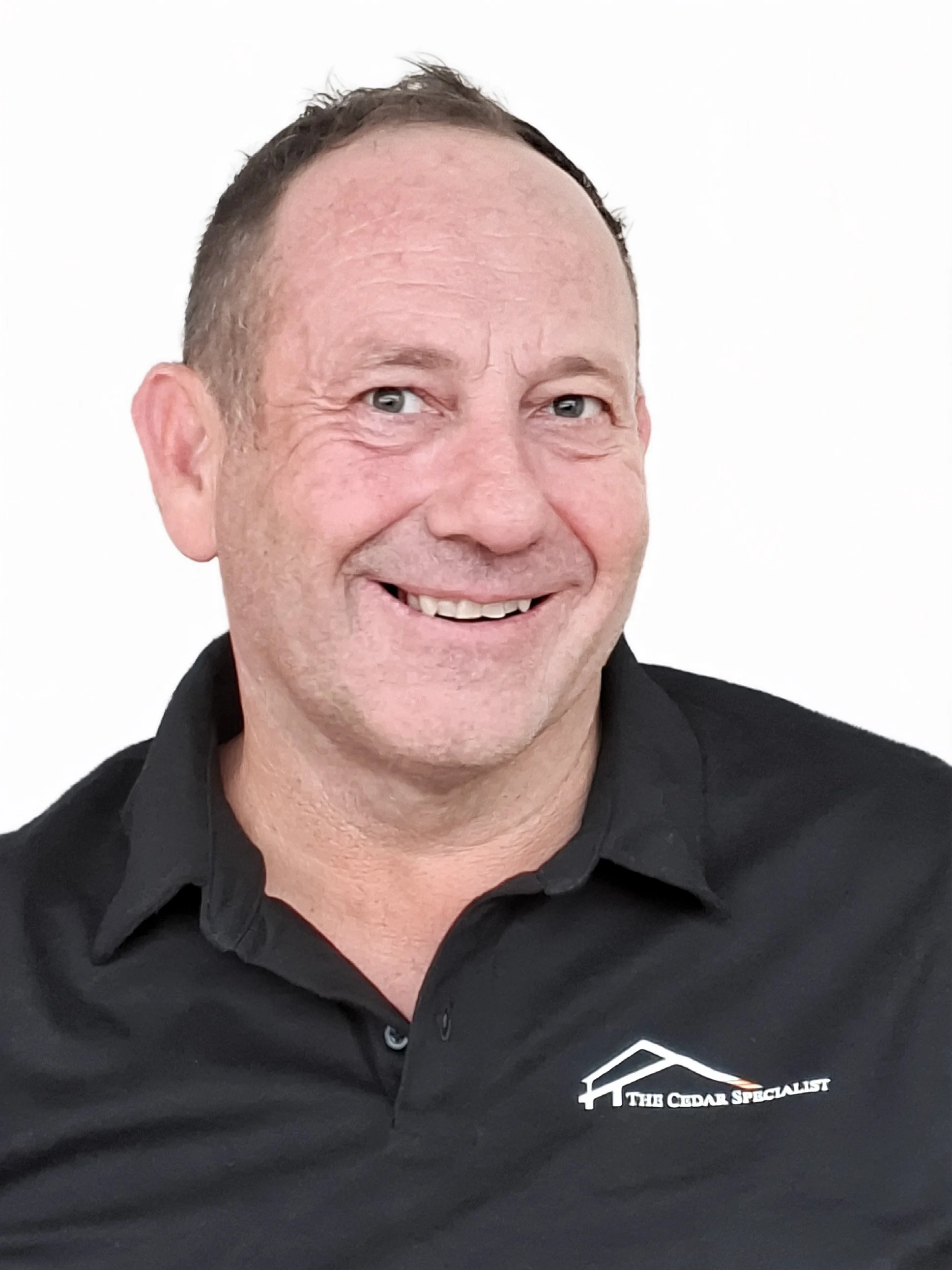 A man smiling, wearing a black collared shirt with a logo of a house roof and the text 'The Cedar Specialist' on it, against a plain white background.
