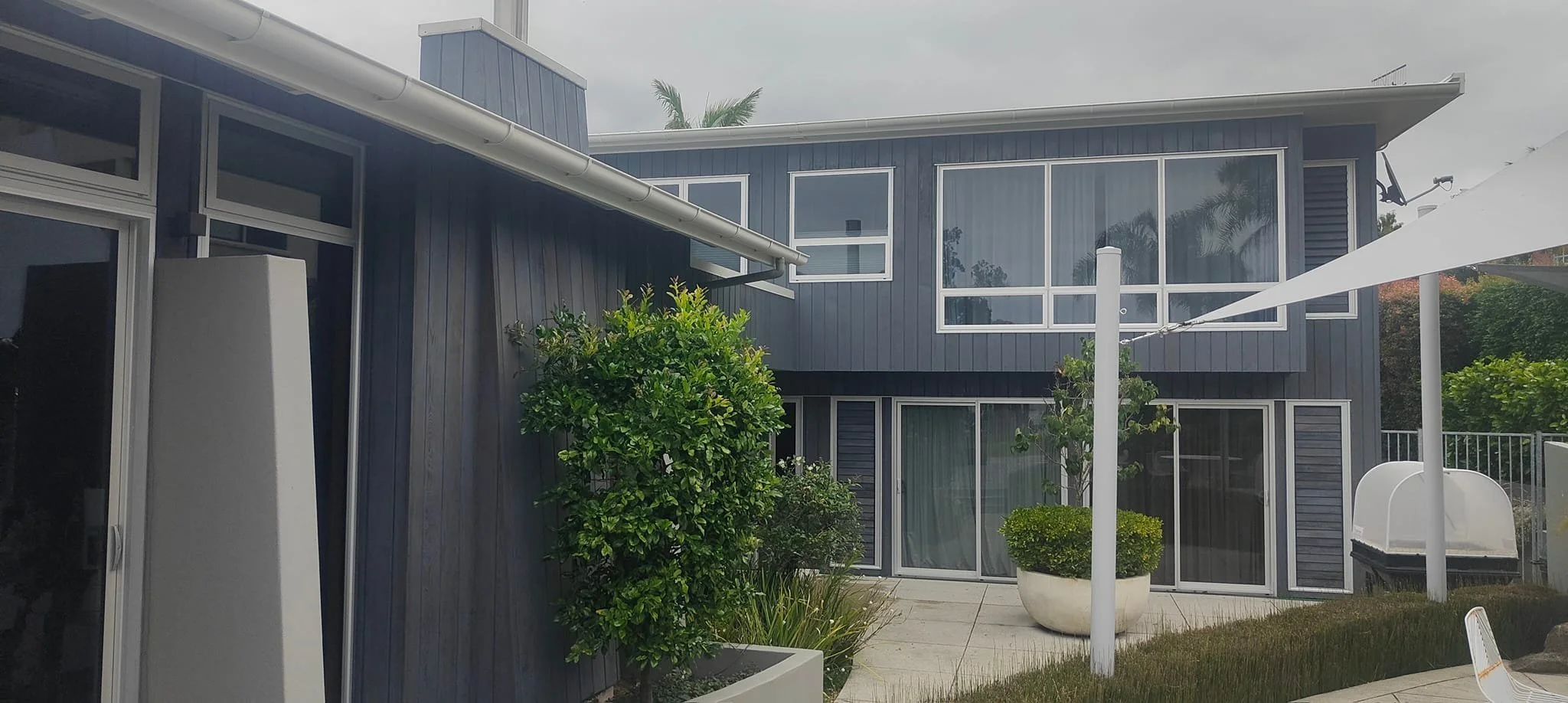 Modern two-story house with large windows, surrounded by greenery, patio with potted plants, and a white outdoor umbrella.