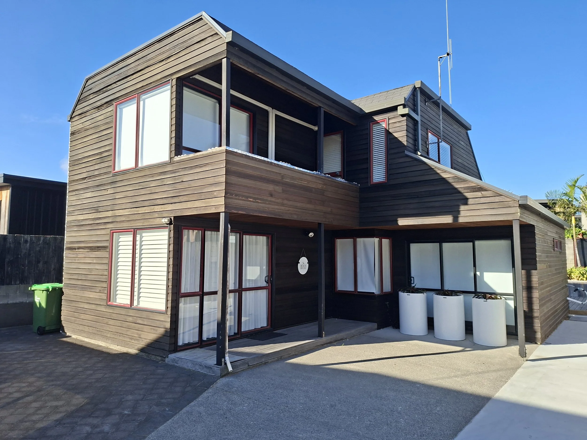 Modern two-story house with dark wooden siding, large windows with red frames, a small balcony, and three white water tanks outside on a concrete pad.