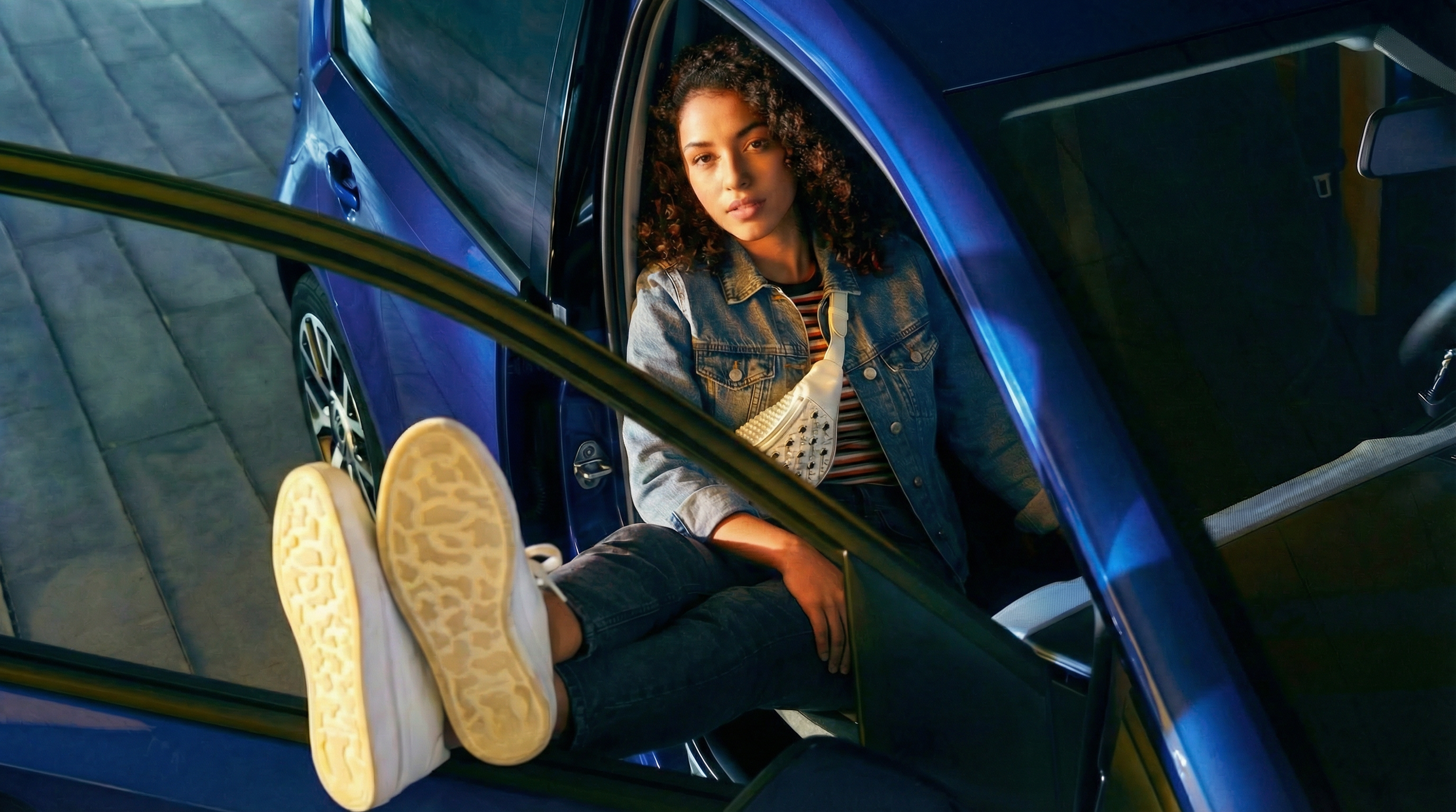 Mujer con cabello rizado, vestida con chaqueta de mezclilla y camiseta a rayas, sentada en un coche azul, viendo hacia arriba, con zapatos deportivos blancos.