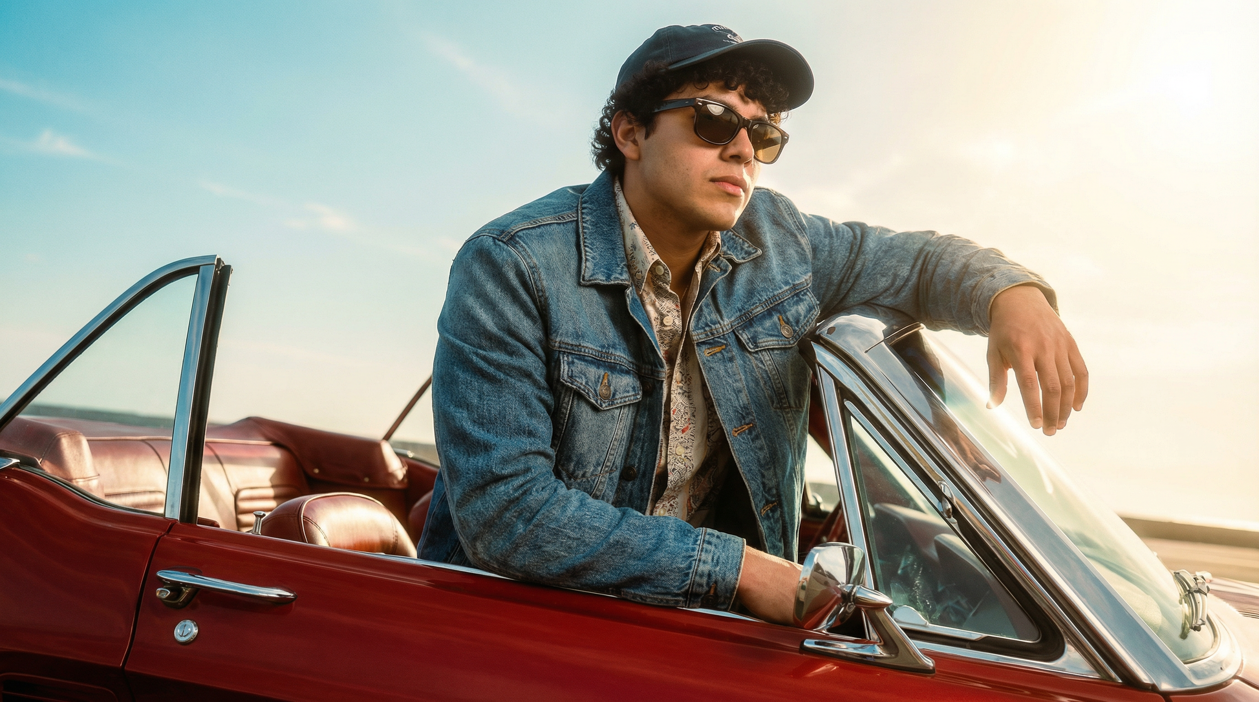 Un joven con gafas de sol, chaqueta de mezclilla y gorra, apoyando el brazo en un coche antiguo de color rojo, en un ambiente al aire libre durante el día.