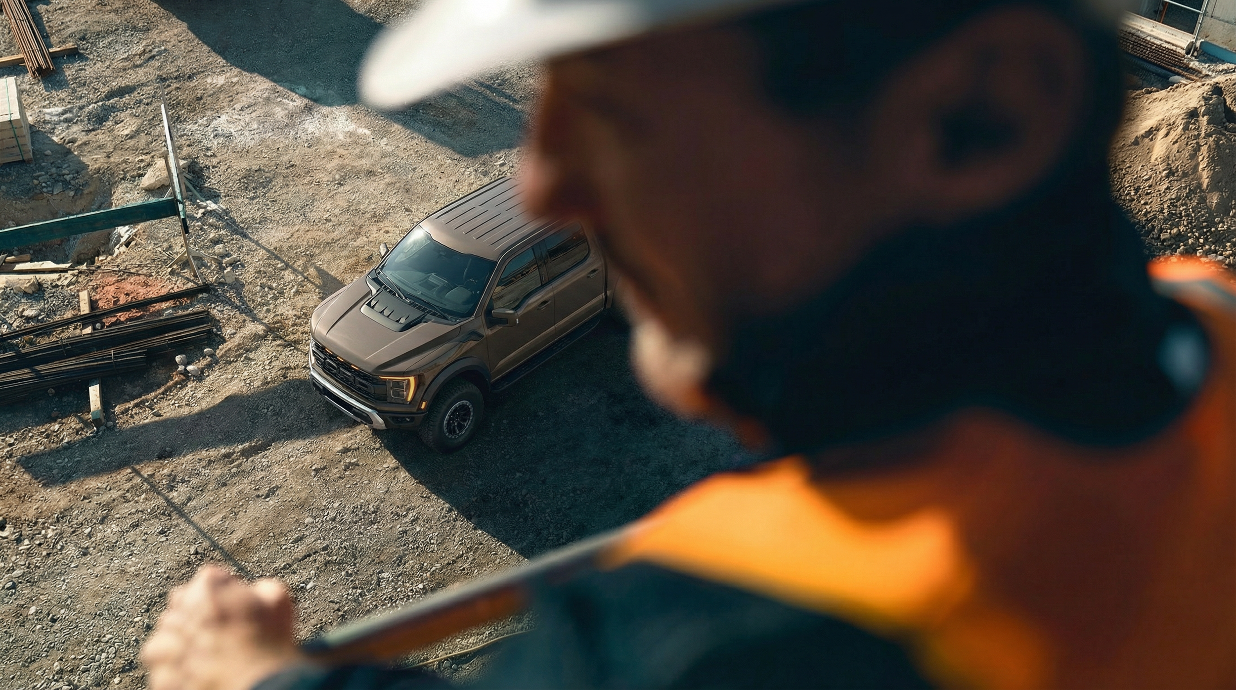 Una persona con casco de seguridad en un sitio de construcción, viendo hacia abajo a un vehículo todoterreno gris en un área de obra con tierra y materiales de construcción.