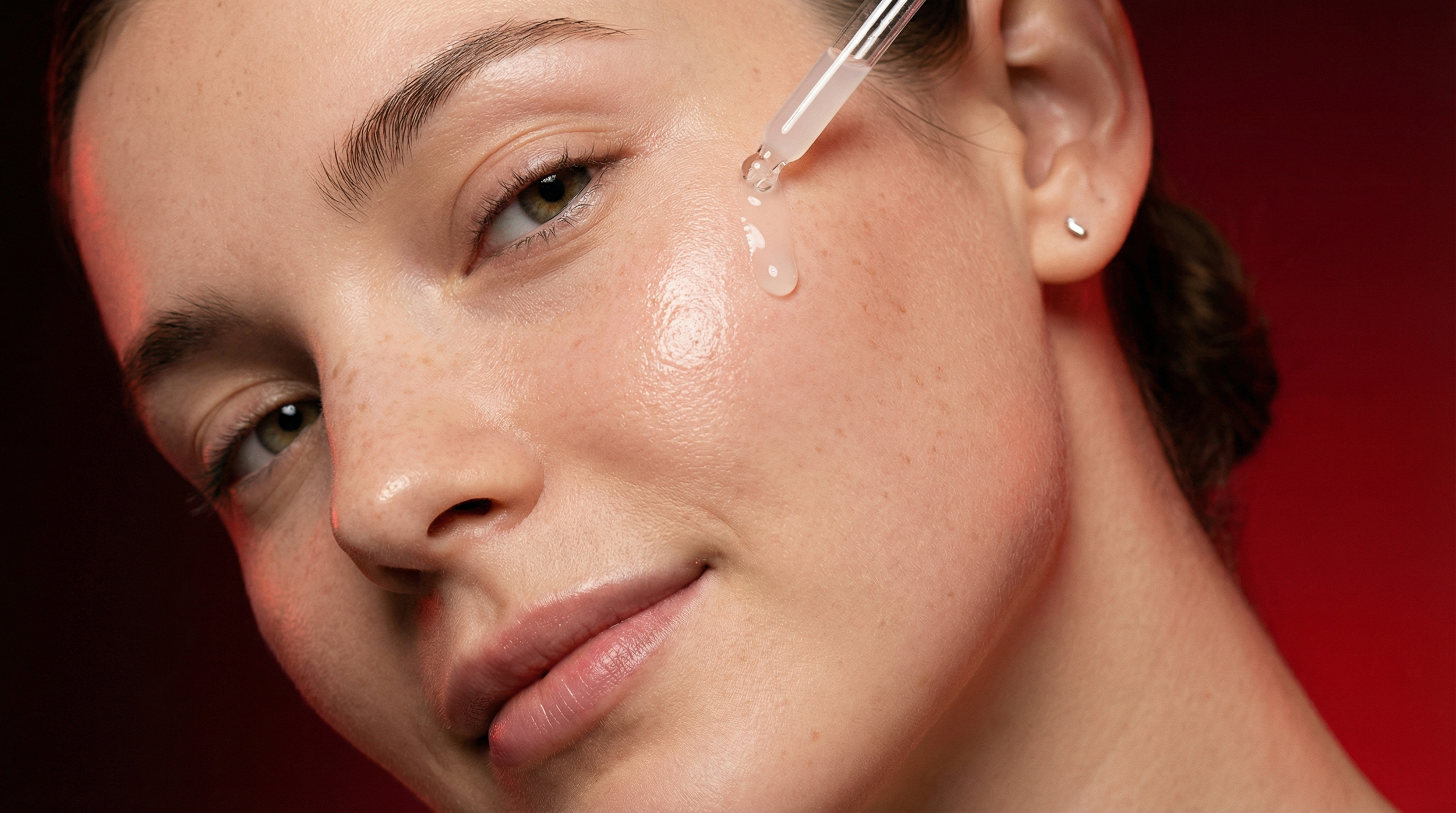 Persona aplicando gotas en la mejilla con un gotero, con piel suave y un fondo rojo.