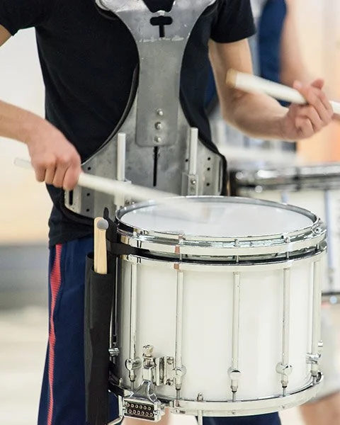 A person playing a snare drum with drumsticks in a marching band uniform.