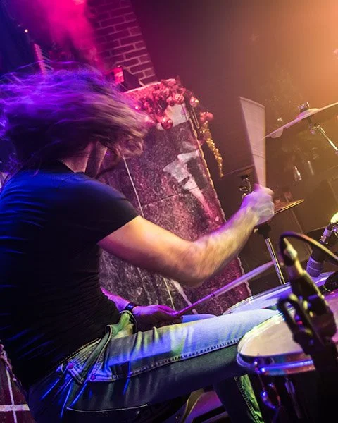 A drummer with long hair playing a drum set on stage, with colorful stage lights, a brick wall, and decorative flowers in the background.