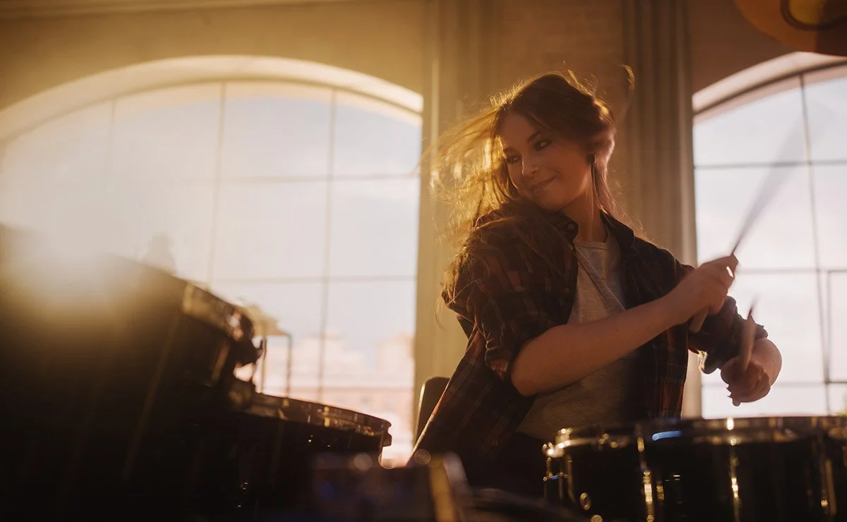 Young woman playing drums in a sunlit room with large windows.