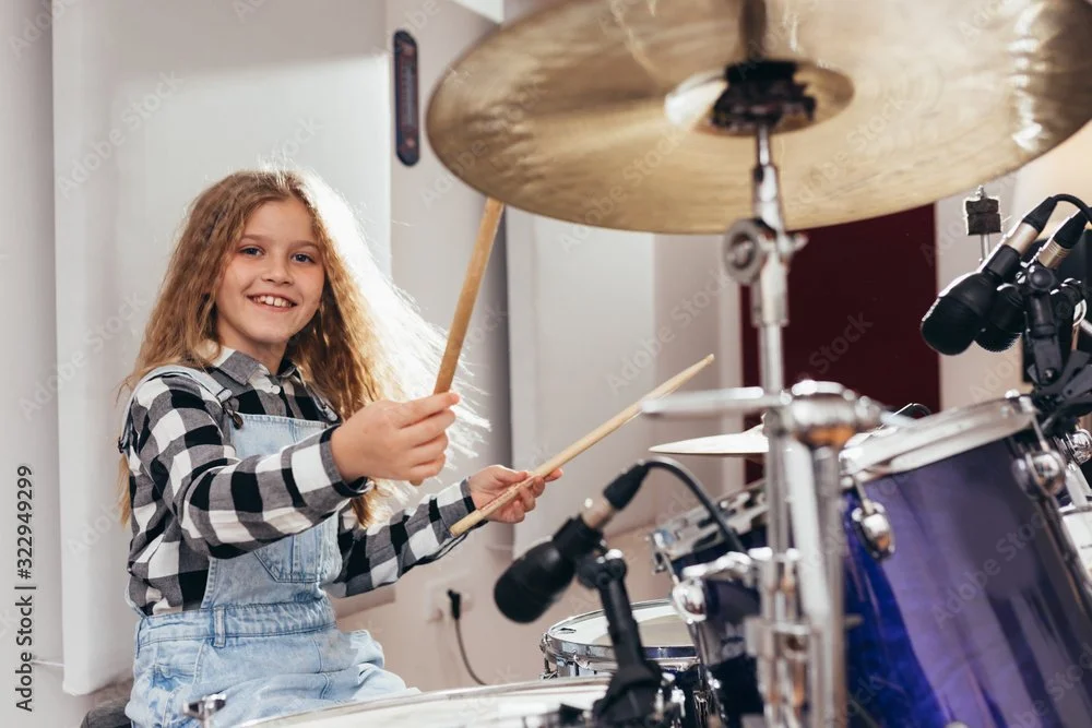 A young girl with long curly hair playing a drum set, smiling, wearing a plaid shirt and denim overalls in a room with sunlight.