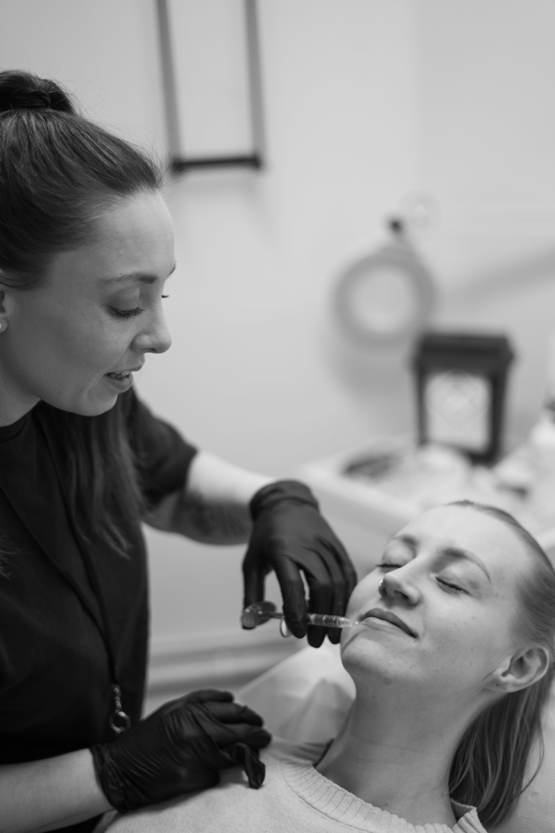 Black and white photograph of an aesthetic nurse practitioner carefully administering a dermal lip filler injection to a relaxed patient in a clinical setting.