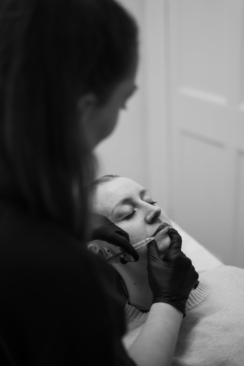 Over-the-shoulder black and white view of an aesthetic nurse wearing black gloves, gently administering a dermal lip filler injection to a relaxed patient.