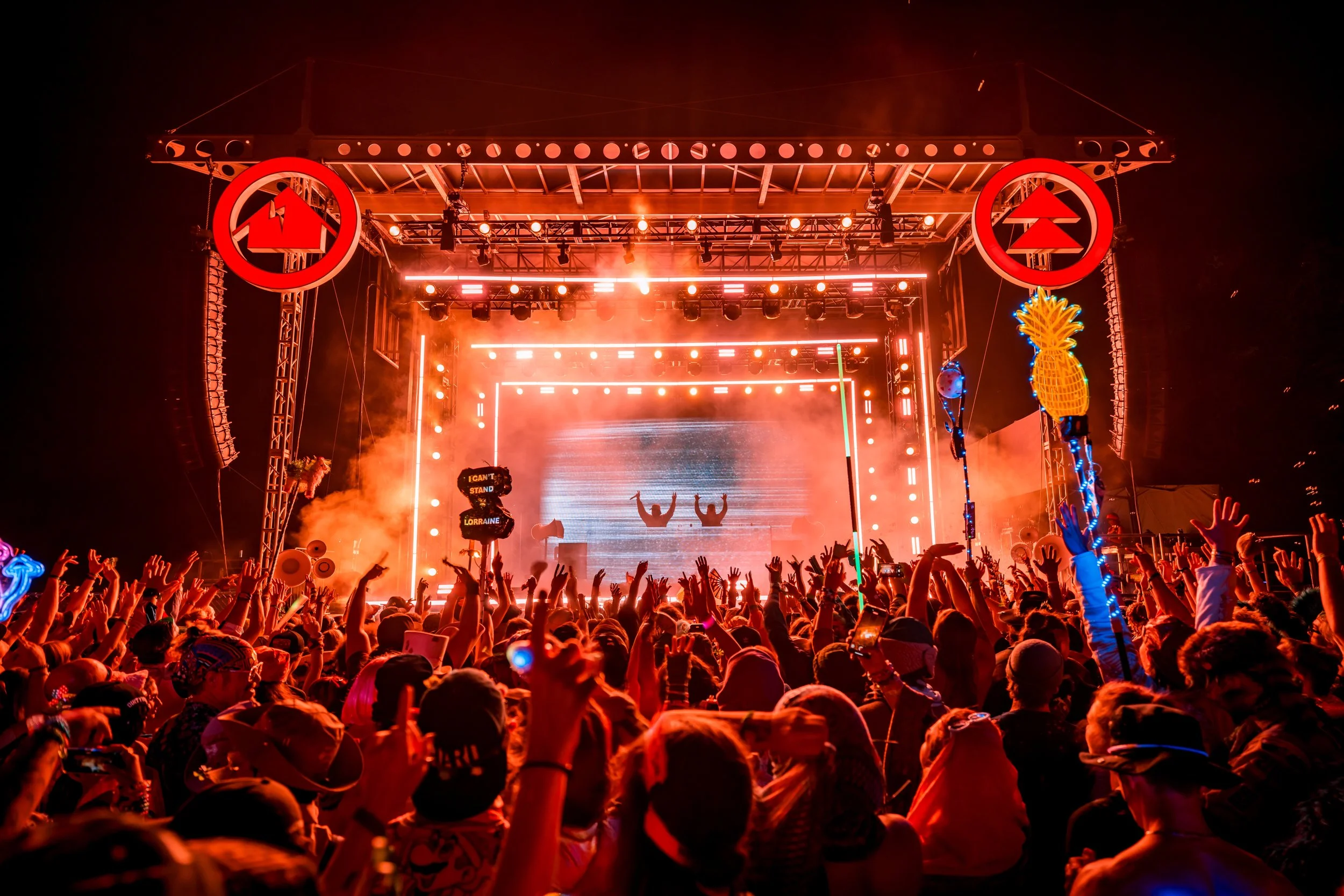 Crowd of people at a music concert with a bright red stage and DJ performing, illuminated with colorful lights and fireworks.
