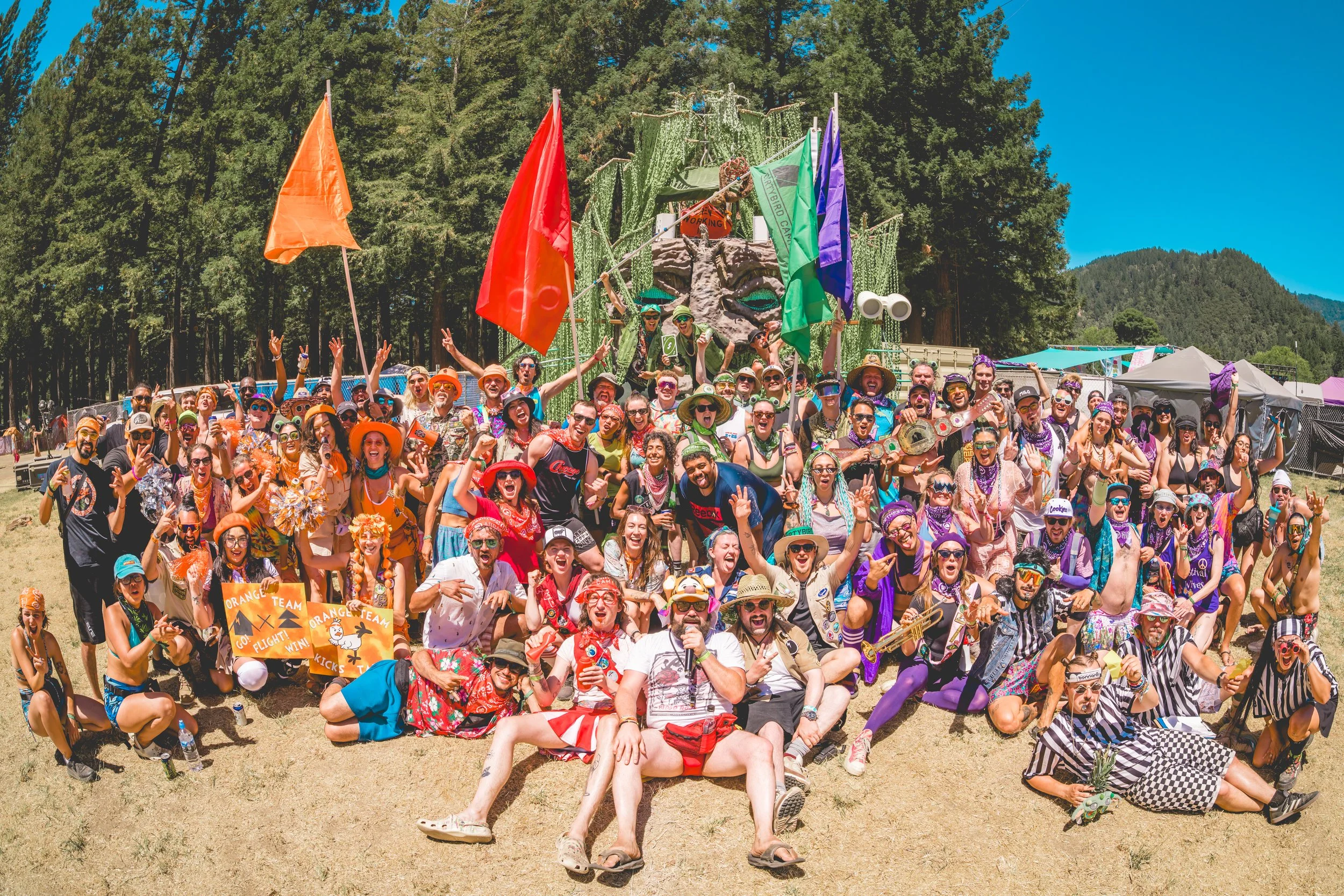 A large group of diverse people gathering outdoors at a festival, smiling and posing for a photo in colorful costumes and accessories, with flags and a decorated structure in the background.