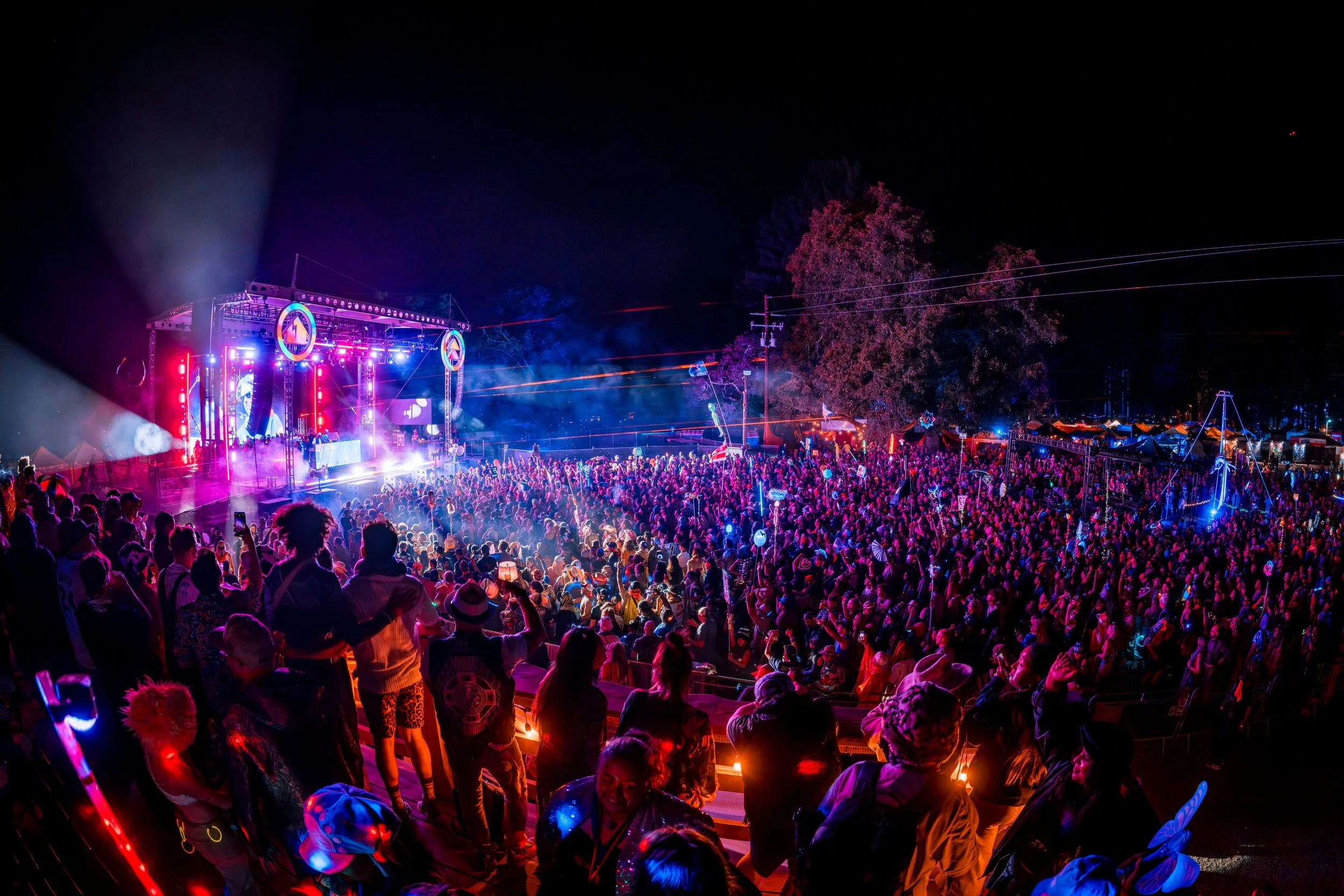 A large outdoor music festival at night with a stage, colorful lights, and a crowd of people dancing and enjoying the concert.