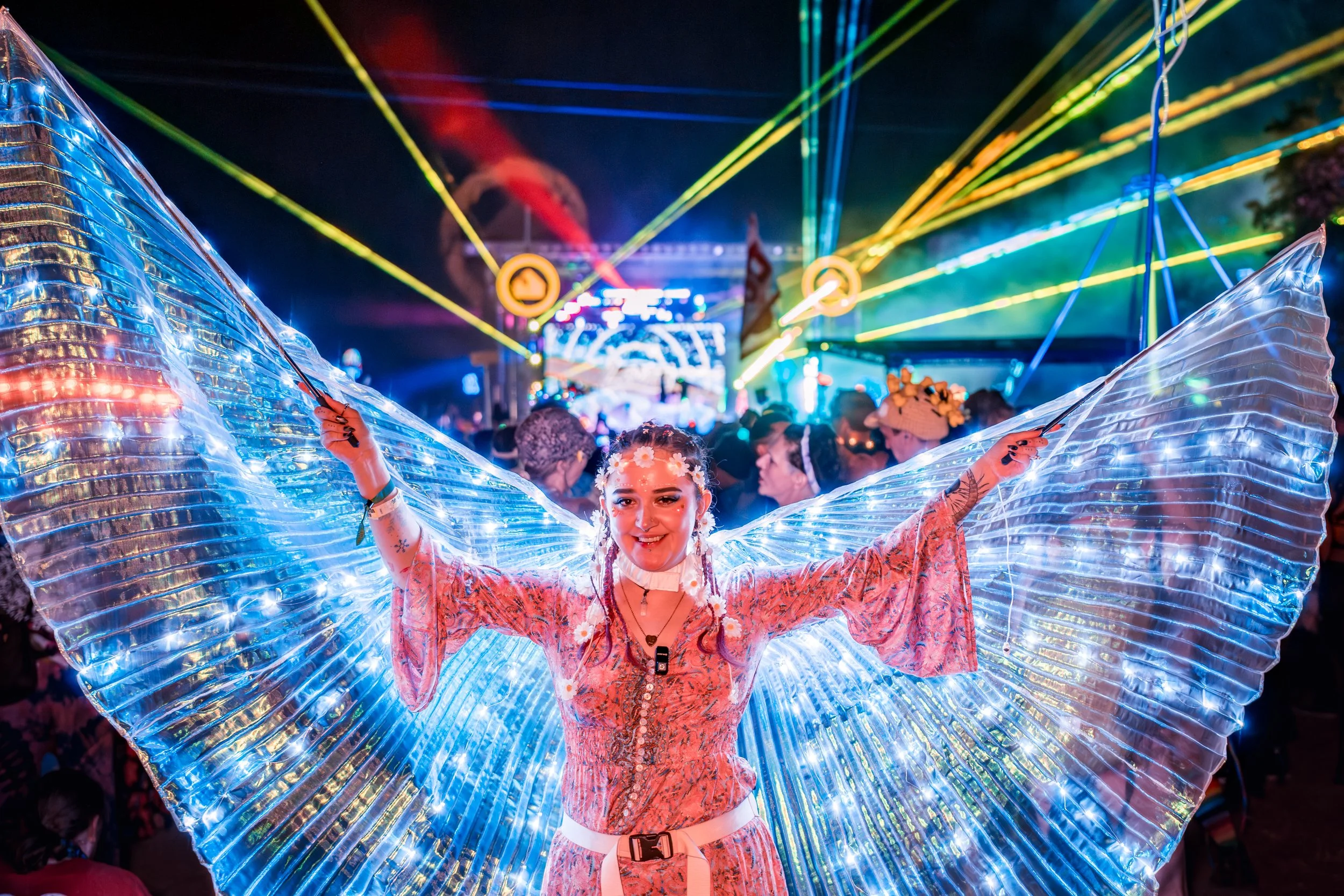 A woman dressed in colorful festival attire, with large, illuminated butterfly wings made of lights, posing with her arms outstretched at a nighttime outdoor music festival with laser lights and a crowd in the background.