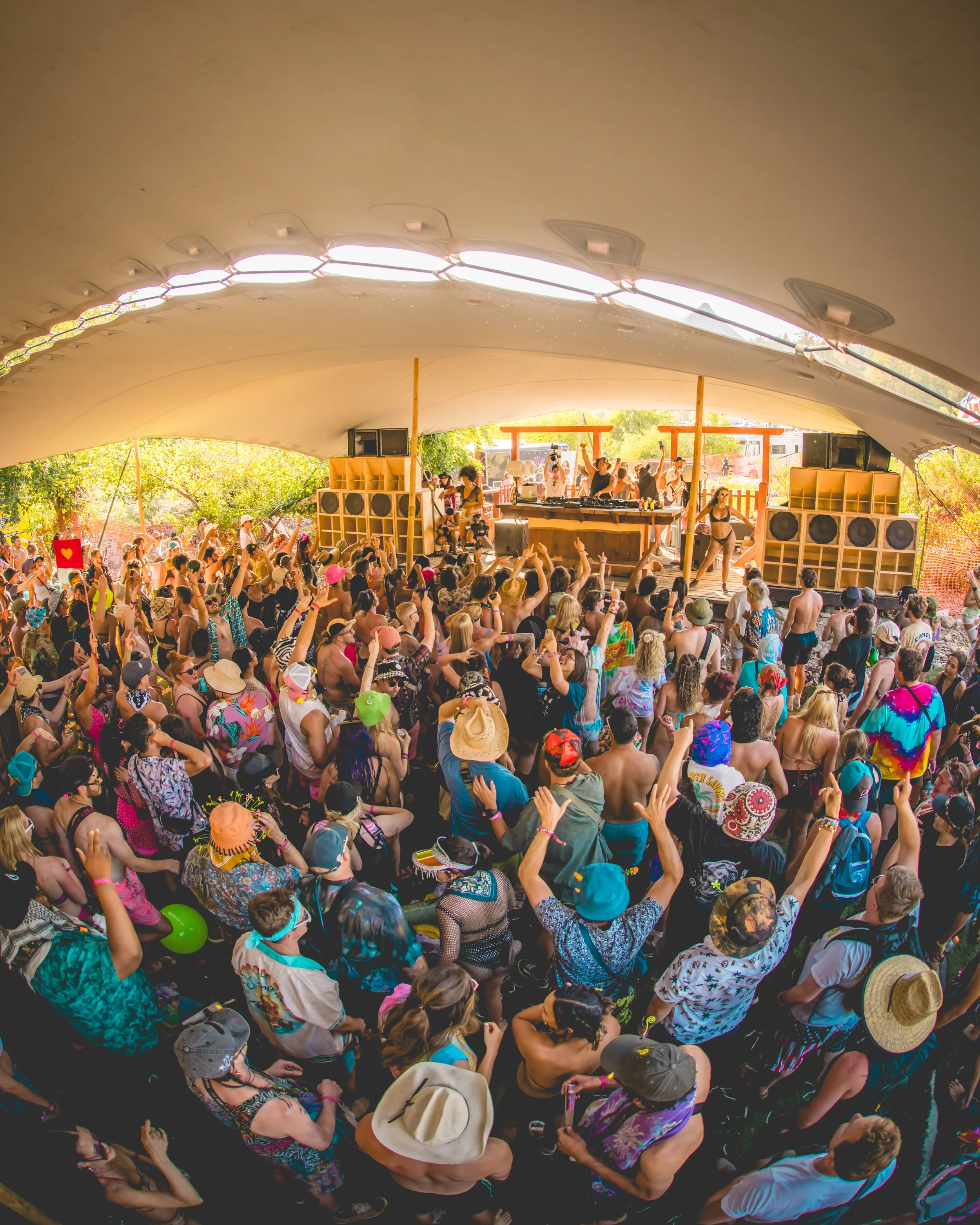 Crowd of people at an outdoor music festival under a large tent, with a stage and DJ booth at the front, decorated with speakers and wooden supports.