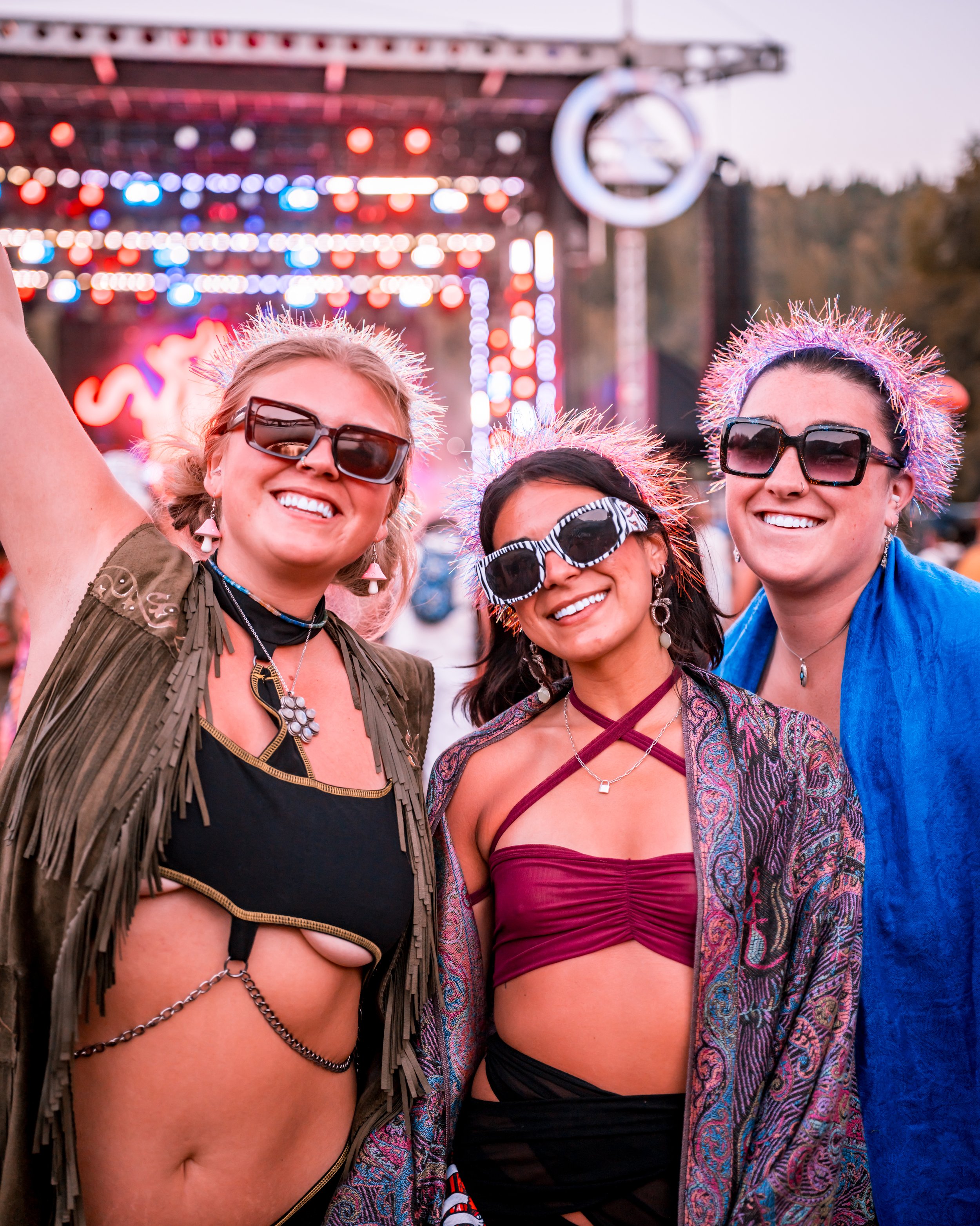 Three people at an outdoor music festival, smiling and wearing sunglasses and colorful clothes with tinsel headbands, with a stage and lights in the background.