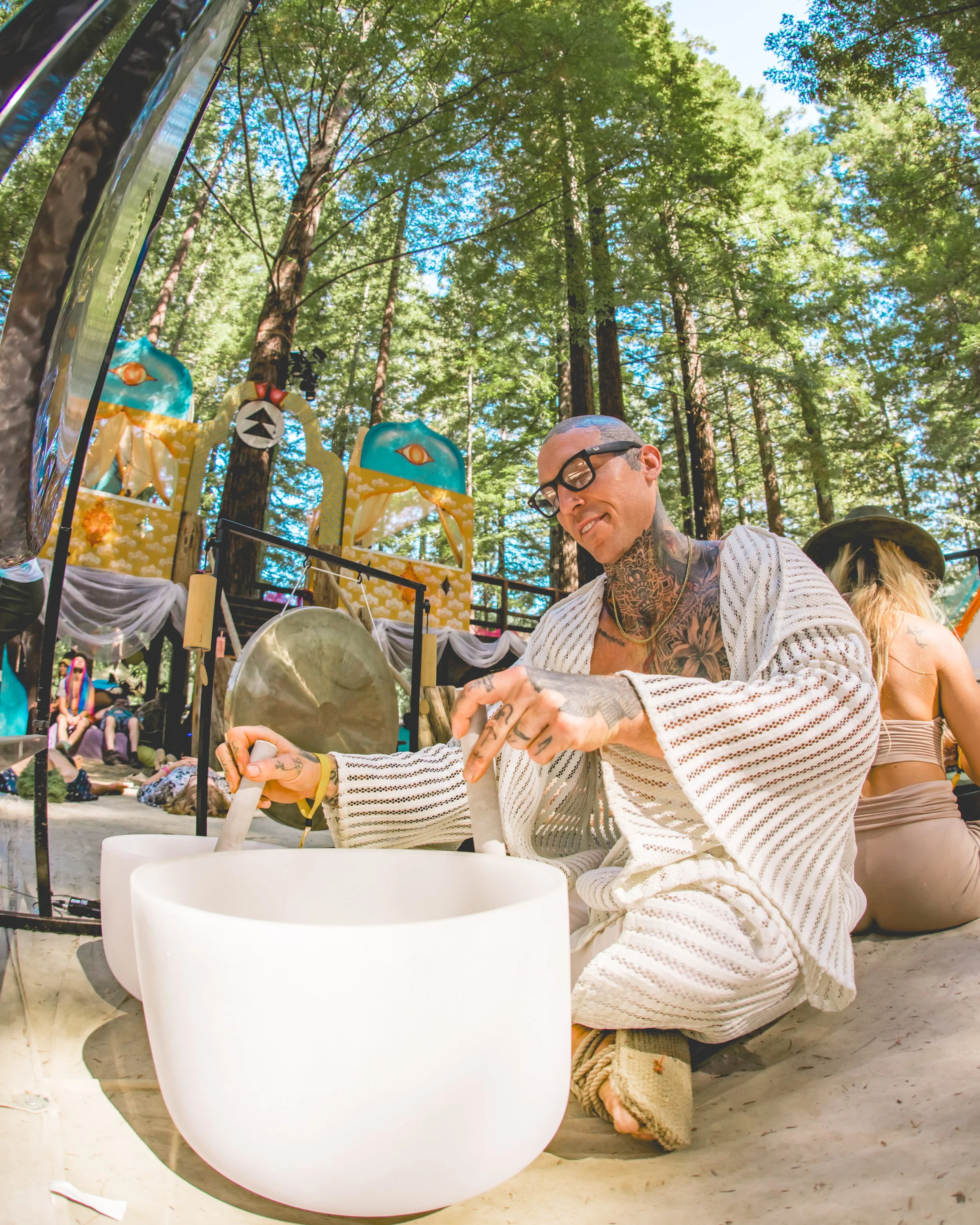 Person with tattoos and glasses, dressed in a striped robe, playing a large singing bowl outdoors at a festival among tall trees, with other festival-goers nearby and colorful decorations in the background.