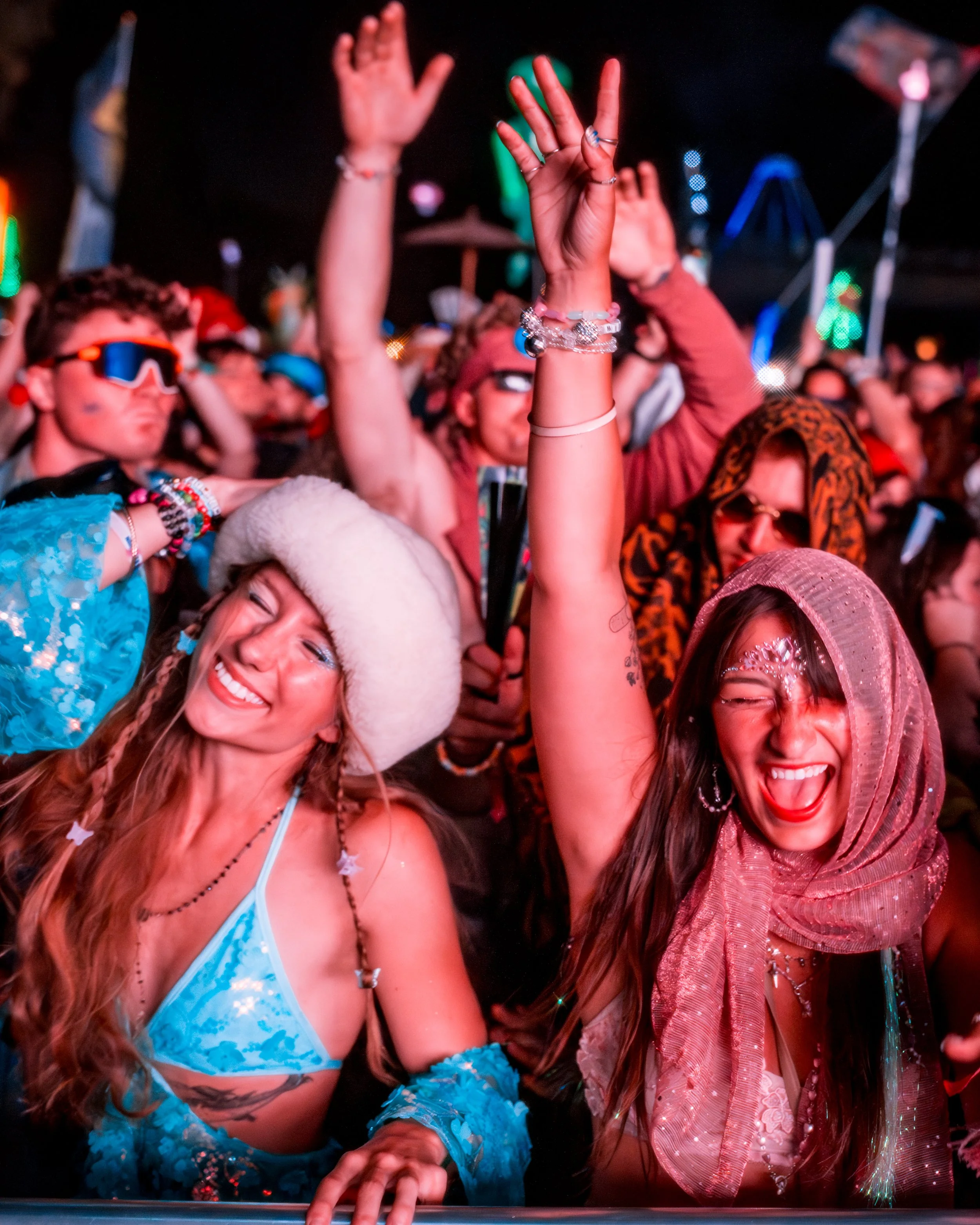 Crowd of festival-goers dancing and enjoying at night, with some raising hands, vibrant lighting, and some wearing colorful and festival-themed clothing and accessories.