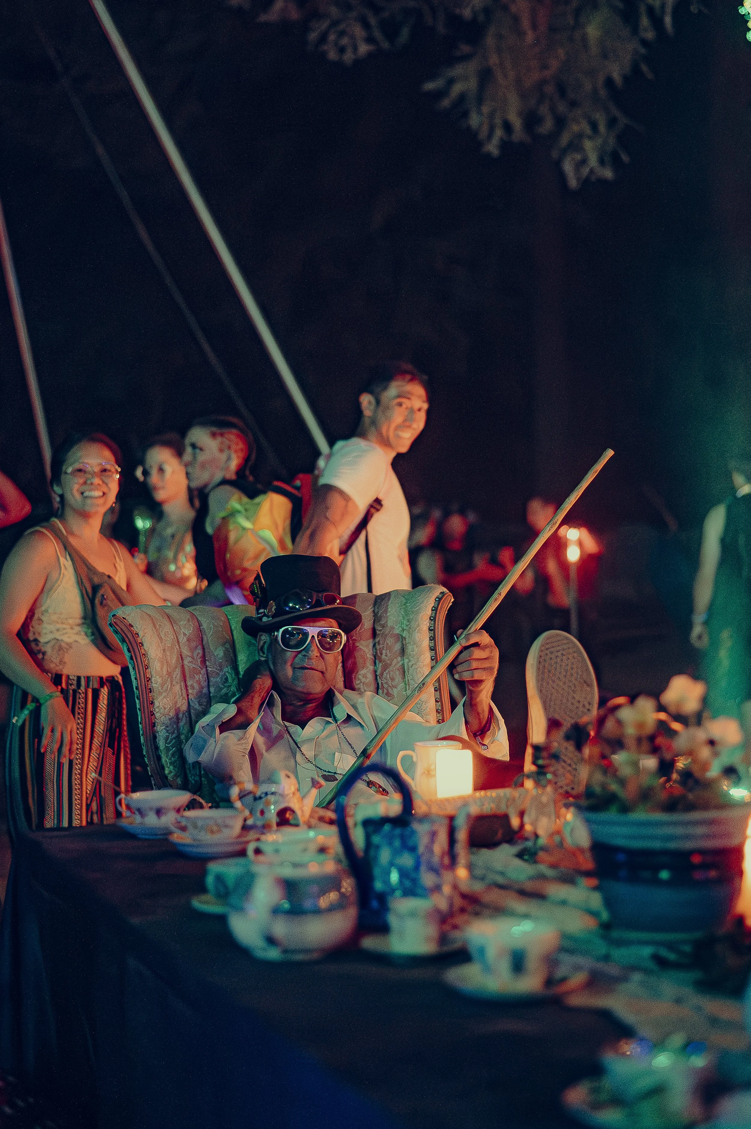 People at a nighttime celebration with an elderly man in front wearing sunglasses and a top hat, sitting in a large armchair with tea sets and food on the table nearby, while others stand behind him smiling and interacting.