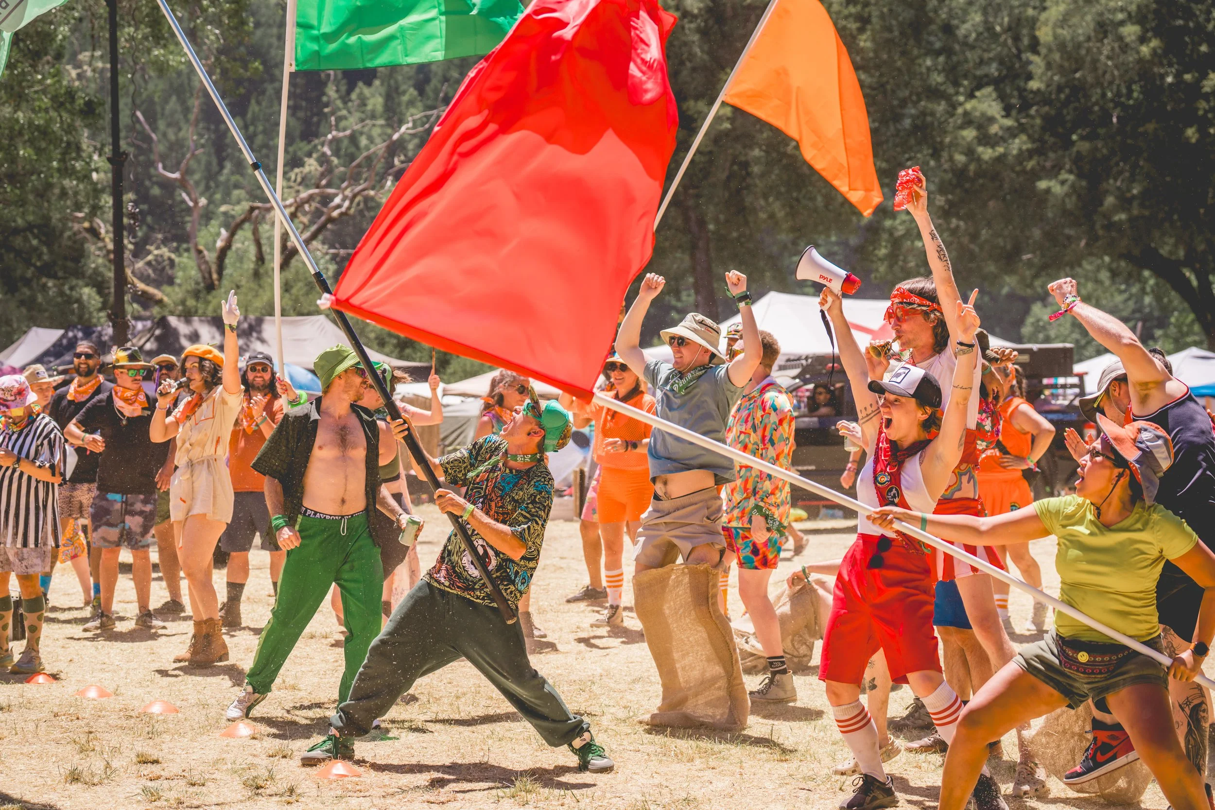 People dancing and celebrating at an outdoor festival, with bright flags, casual summer clothing, and some wearing sunglasses and hats.