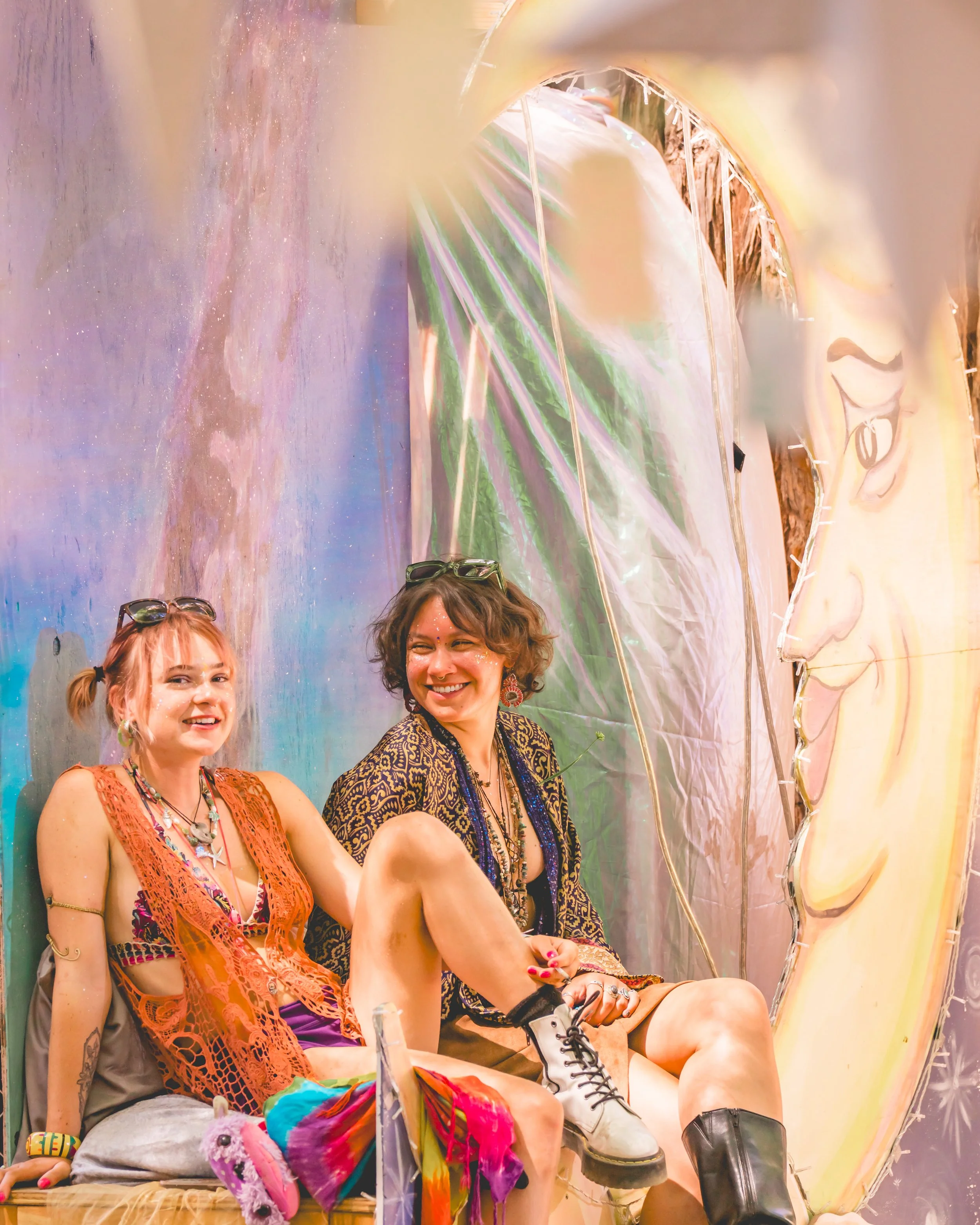 Two women sitting on a colorful bench in front of large, whimsical art featuring a face and vibrant patterns; one woman has reddish hair, and the other has brown hair; both are wearing casual, eclectic clothing with accessories.