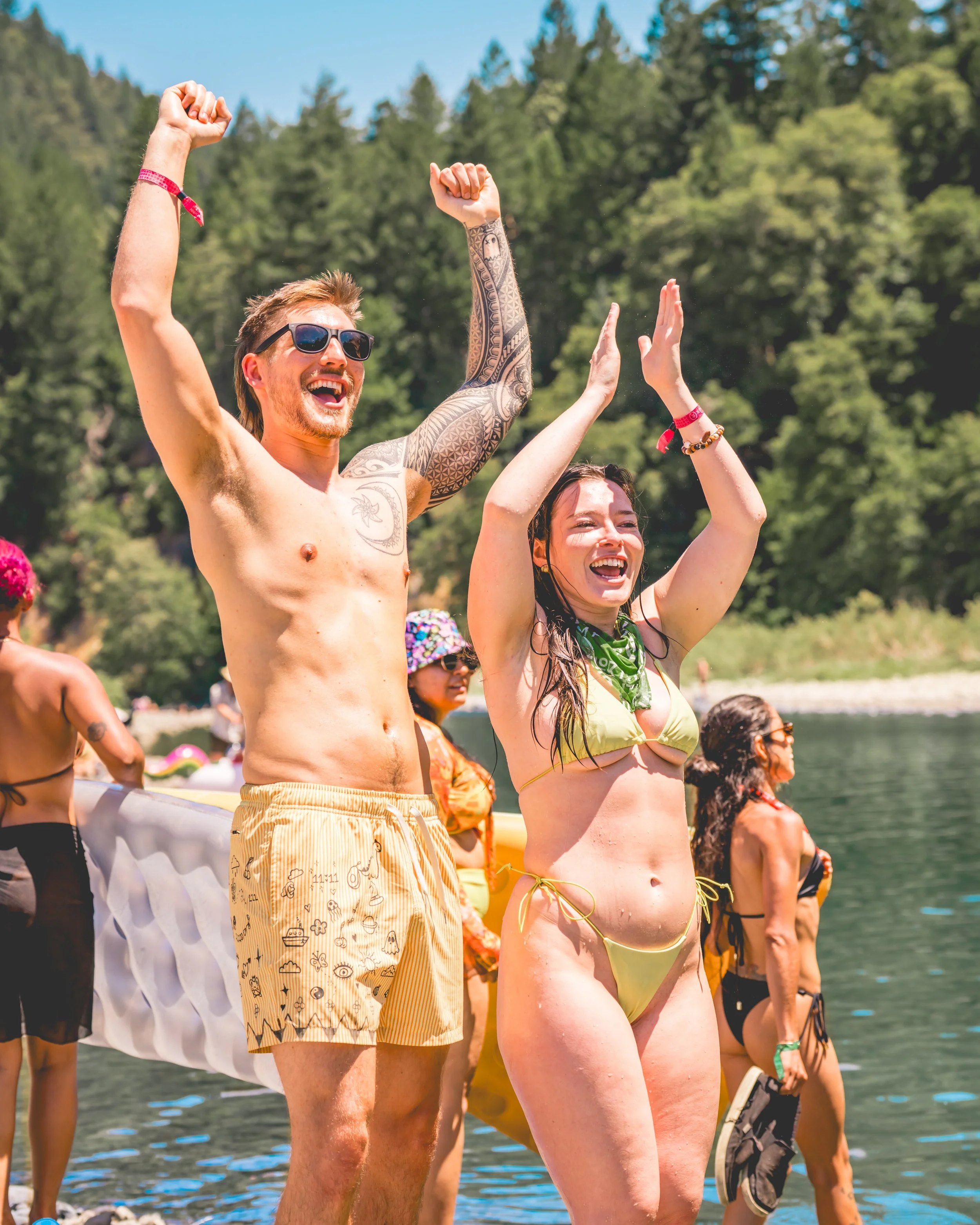 People celebrating at the lake, with their arms raised, enjoying sunny weather and surrounded by trees.