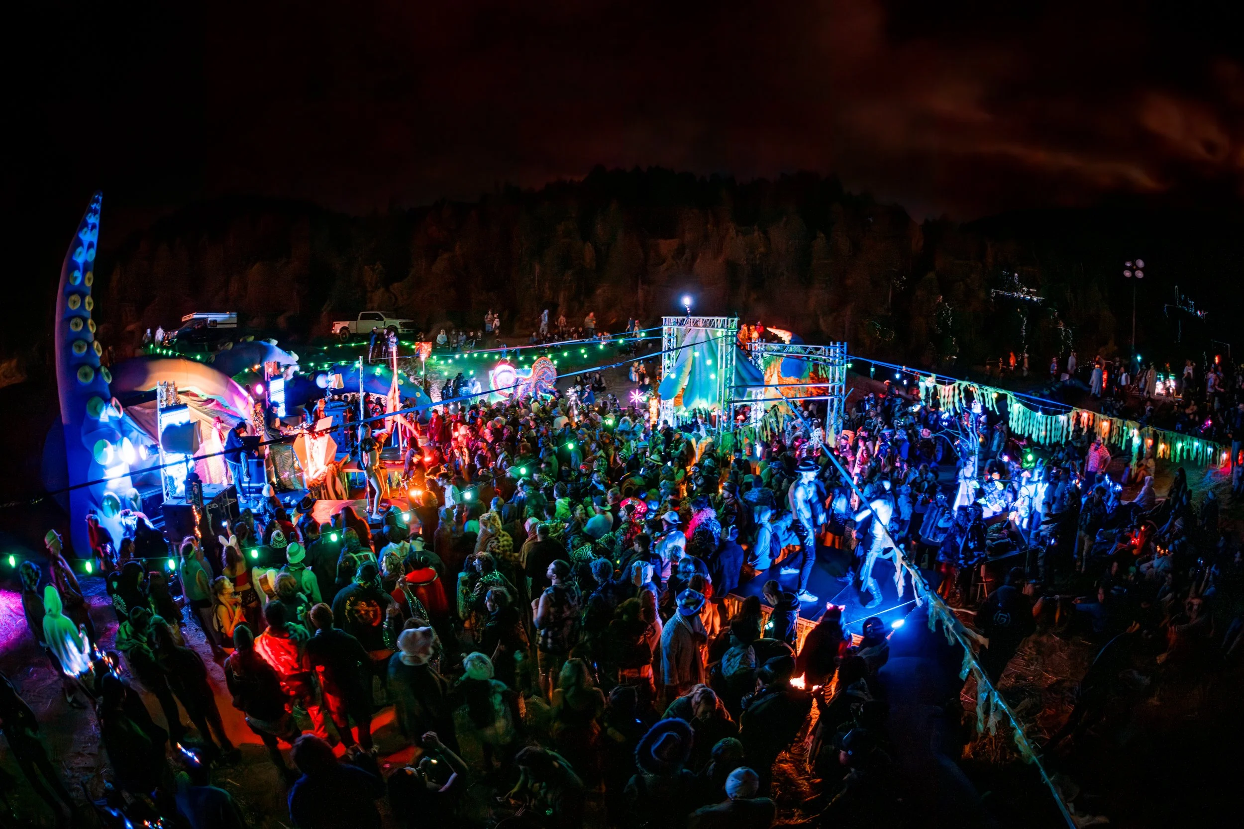 A large outdoor nighttime festival or concert with a crowd of people, colorful lights, and stage decorations set against a dark sky and rocky landscape.