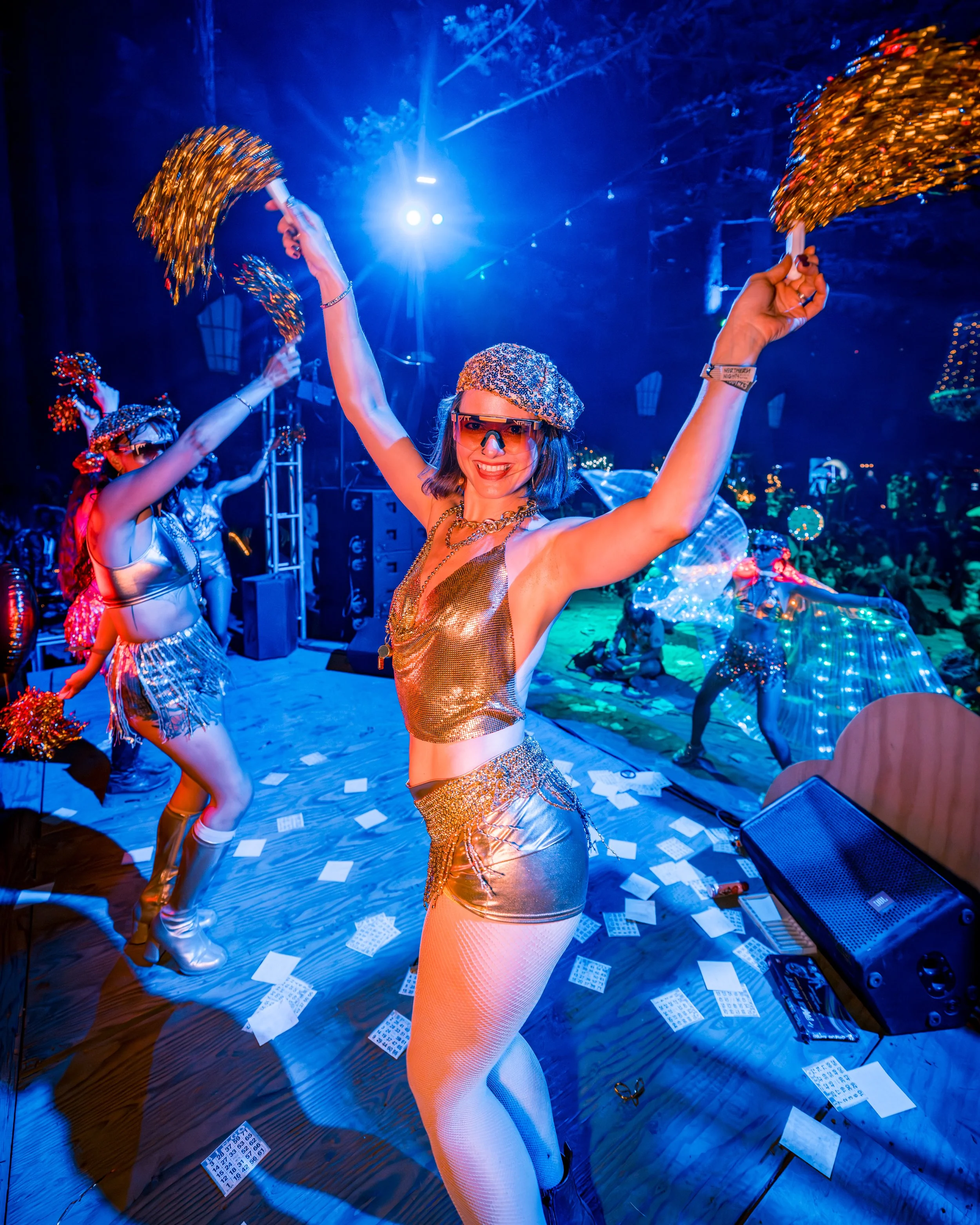 Women dancing on stage at a party with shiny outfits, pom-poms, and colorful lighting, surrounded by scattered cards on the floor.