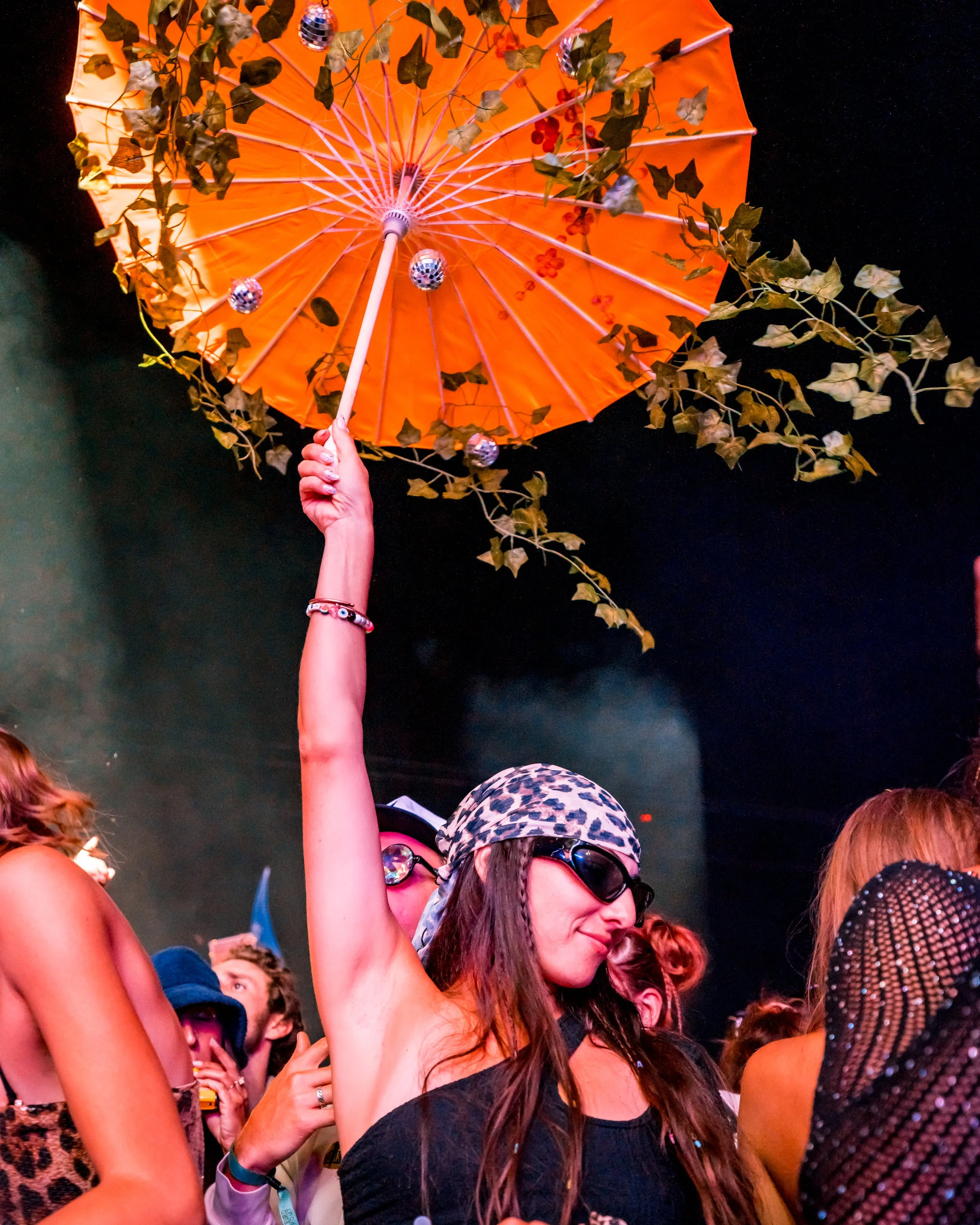 A woman holding an orange umbrella with leaves and small disco balls inside, wearing a leopard print headscarf and sunglasses at a party or concert.