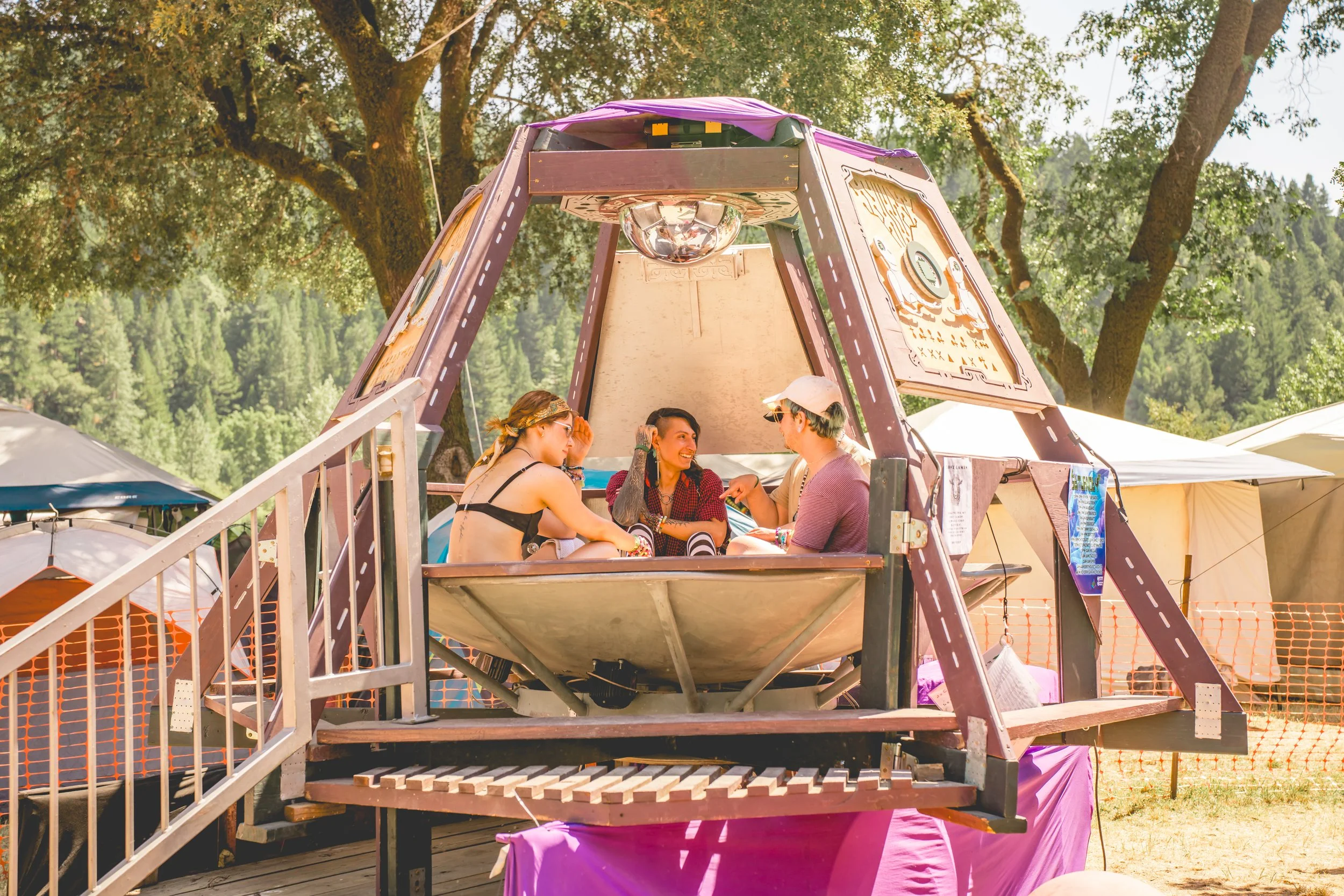 Three people sitting inside a purple and wooden observational structure at an outdoor festival during daytime.