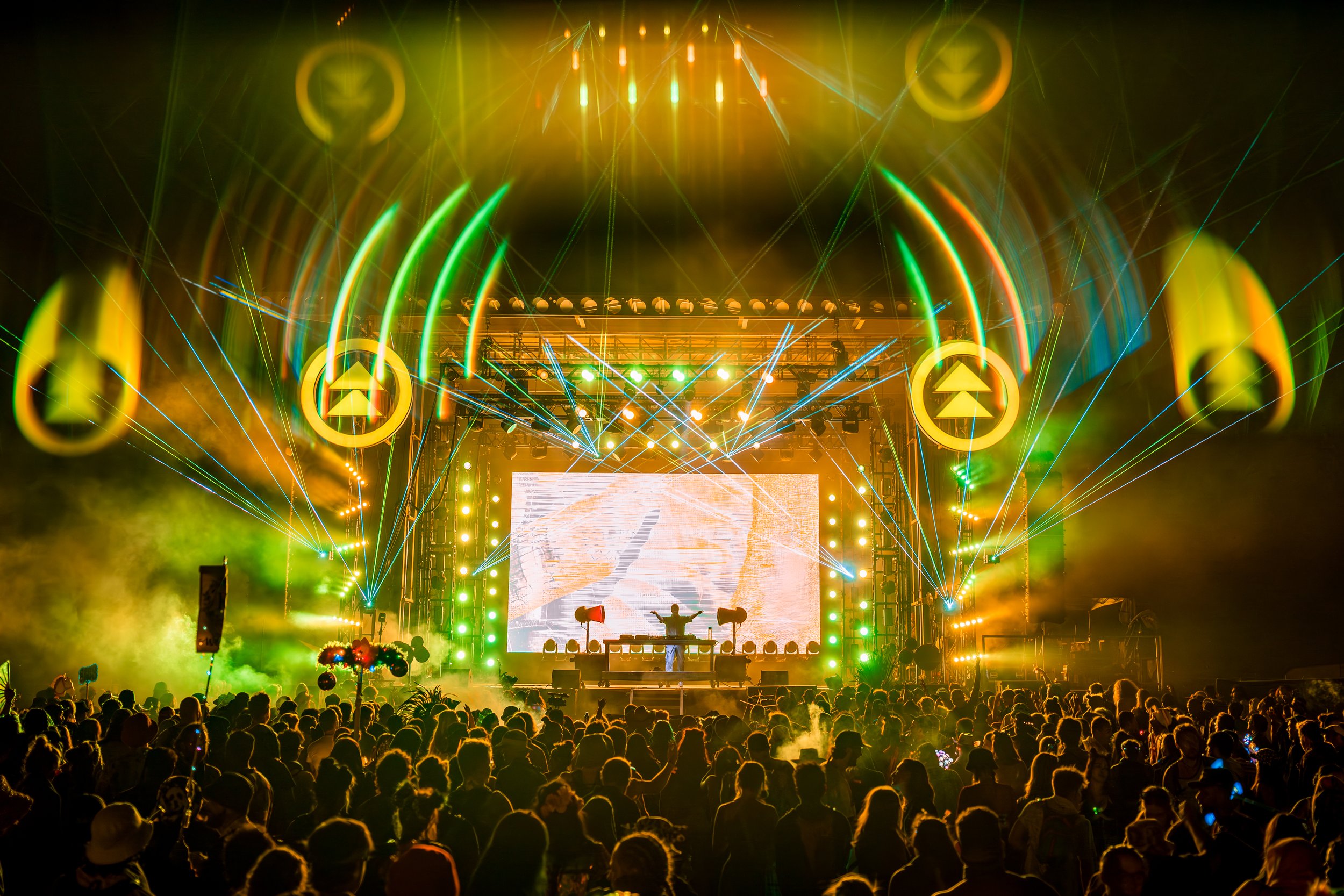 A vibrant concert scene with a large crowd, colorful stage lighting, lasers, and a DJ performance on stage with a large digital screen in the background.