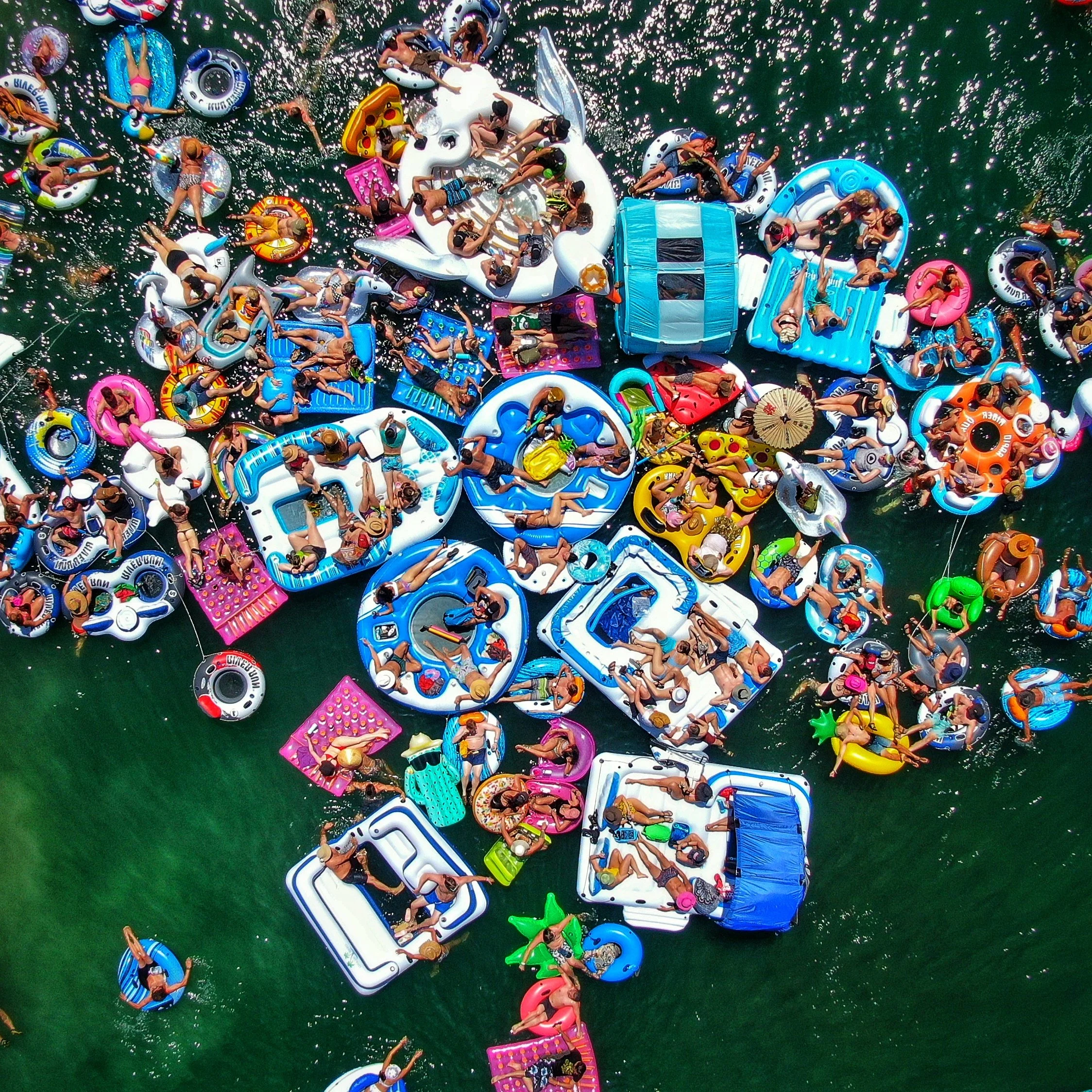 Aerial view of many people on colorful inflatable pool floats and boats in a body of water, enjoying a sunny day.