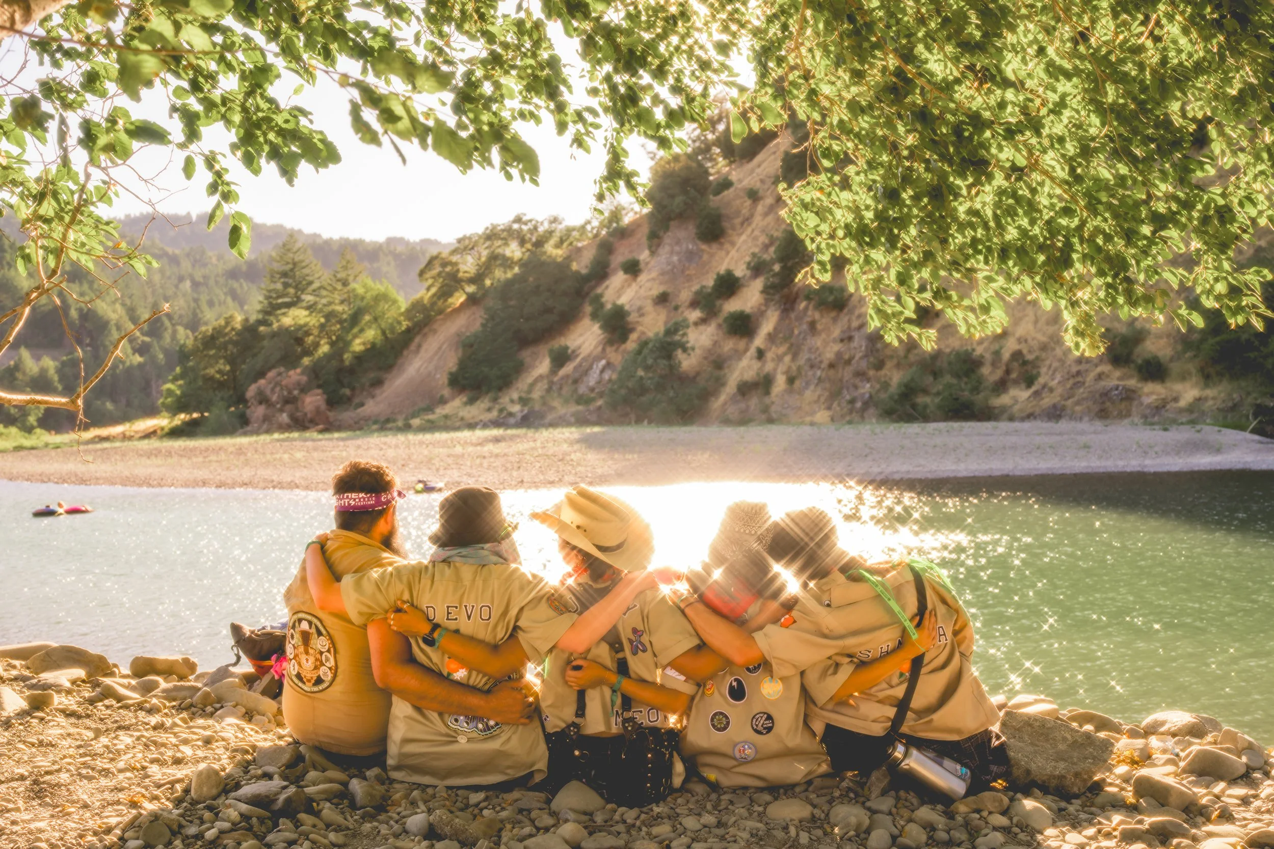A group of five scouts sitting on a rocky riverbank with their arms around each other, overlooking a body of water with hills in the background, and the sun shining brightly through tree leaves.
