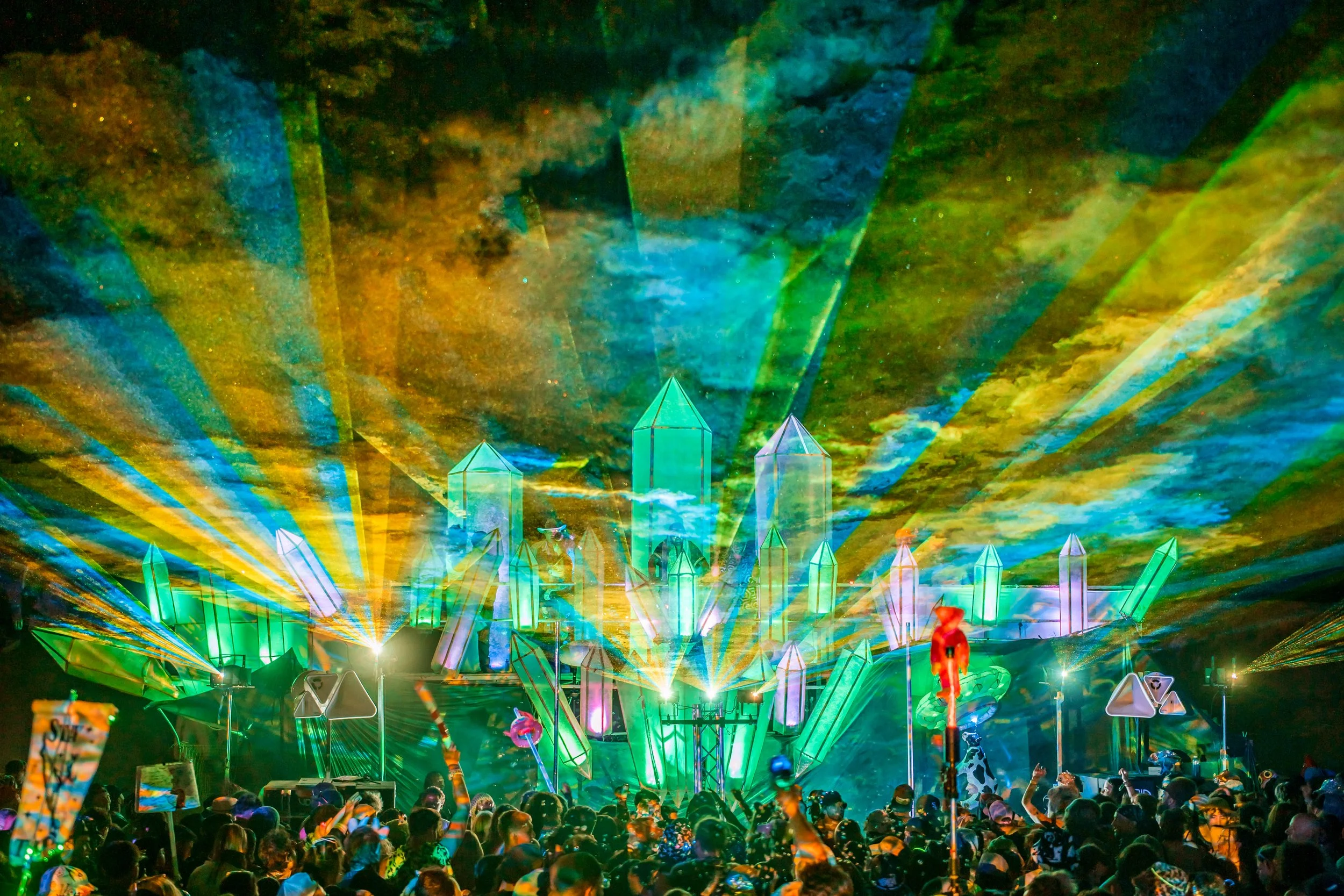 A vibrant concert scene with a colorful, laser-lit stage featuring geometric structures and a crowd of people enjoying the event.