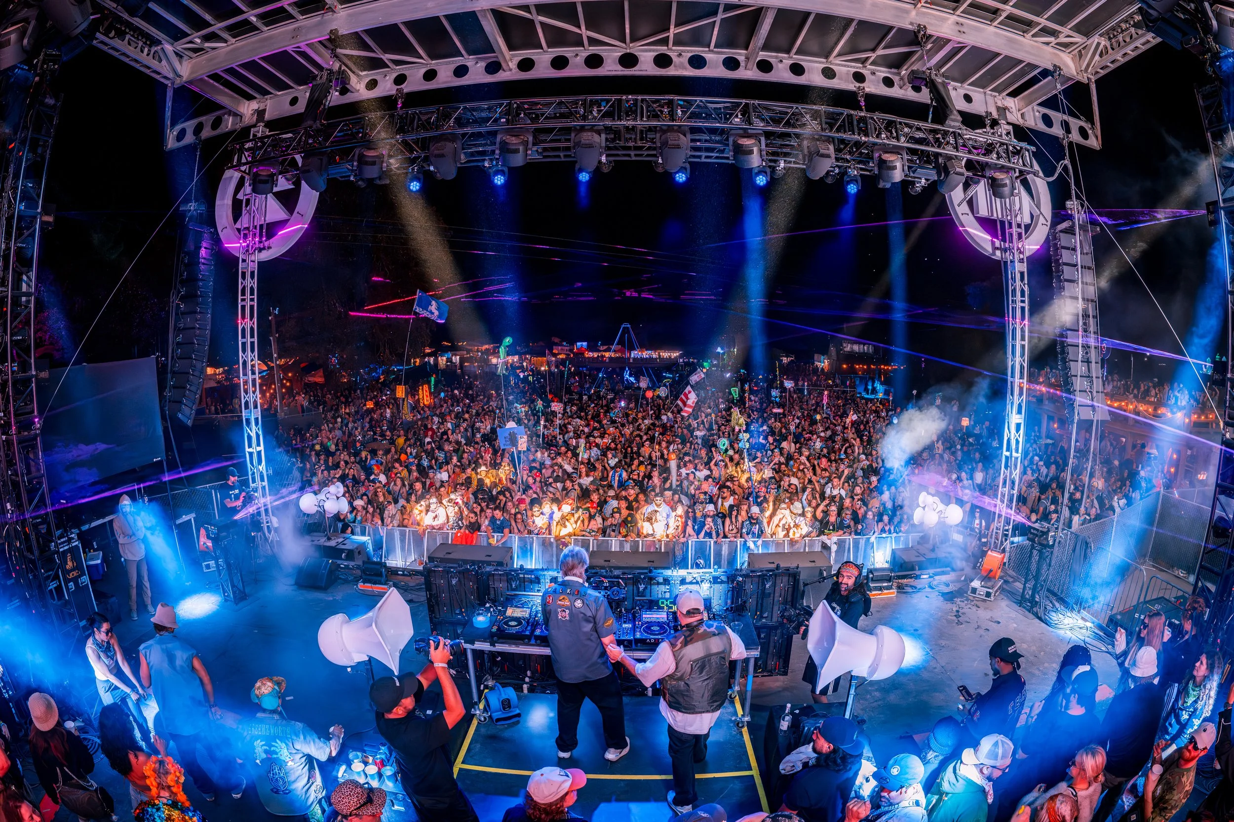 A large outdoor music festival at night with a crowd of people on the main stage, DJ equipment, colorful lights, and fog effects.