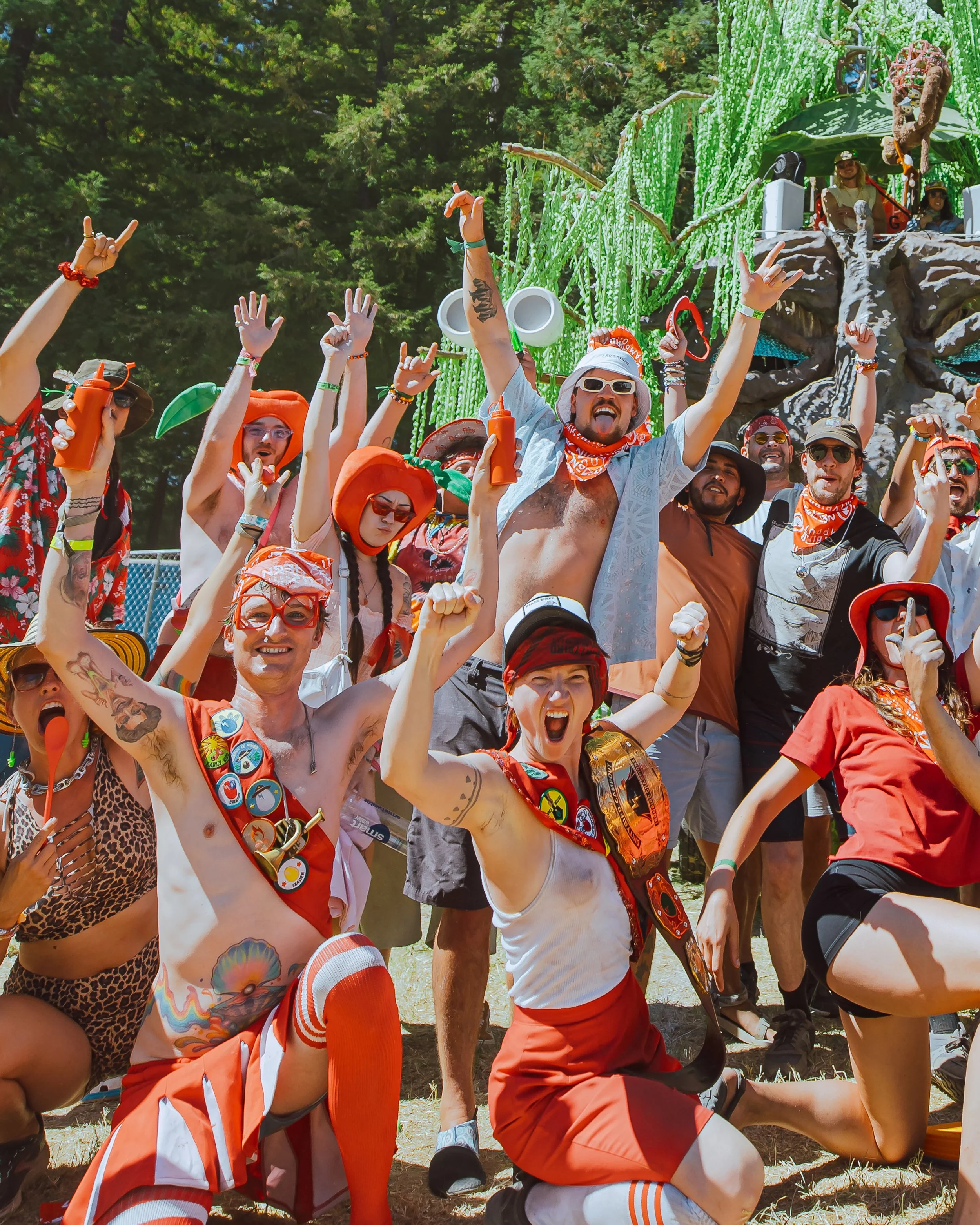 A group of people at an outdoor festival celebrating with excitement. They are dressed in colorful, festive clothing and accessories, some with face paint, hats, and sunglasses, raising their arms in celebration. In the background, there is a large d