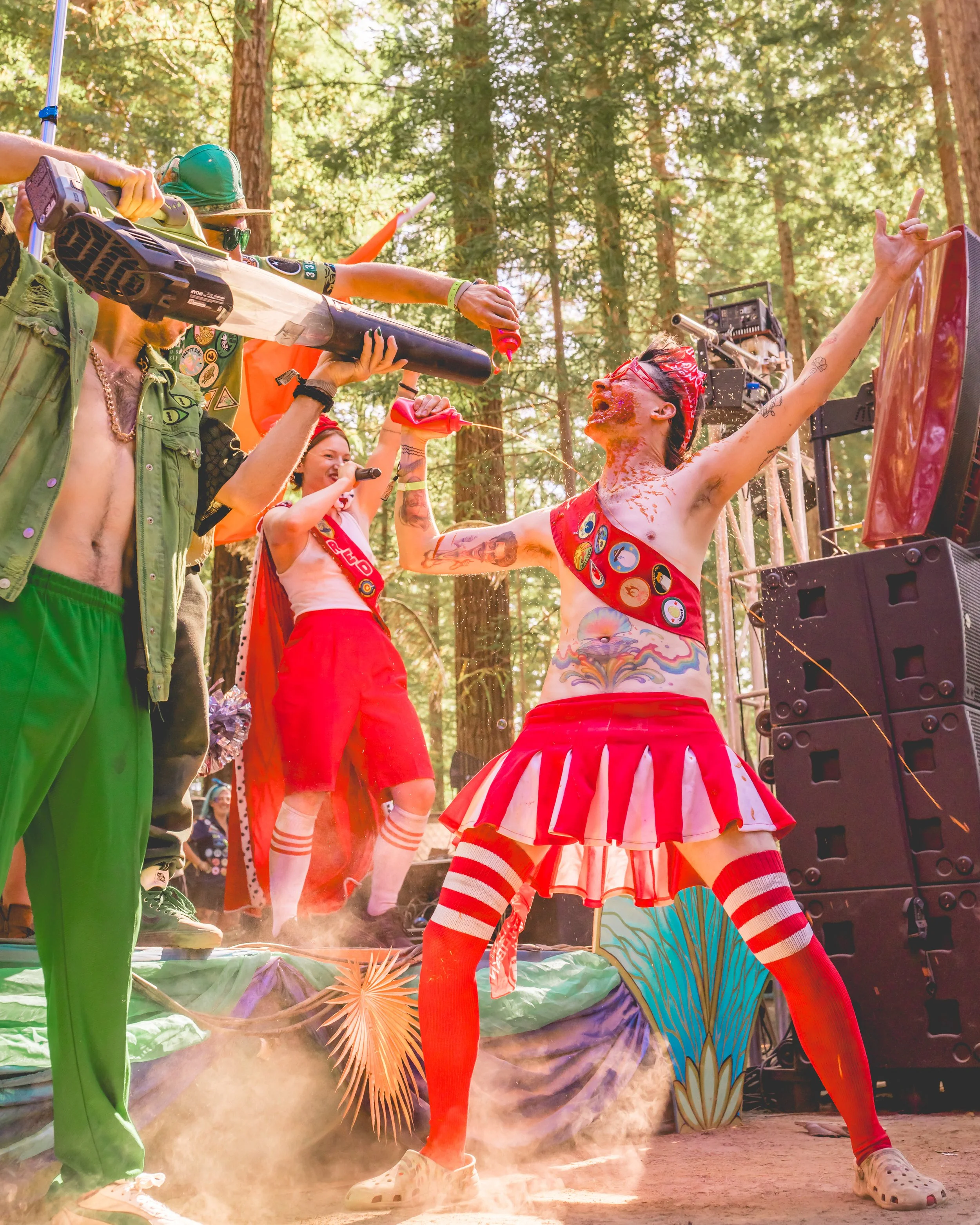 People in colorful costumes spray water, play, and celebrate at an outdoor event in a forested area, with trees and stage equipment visible.