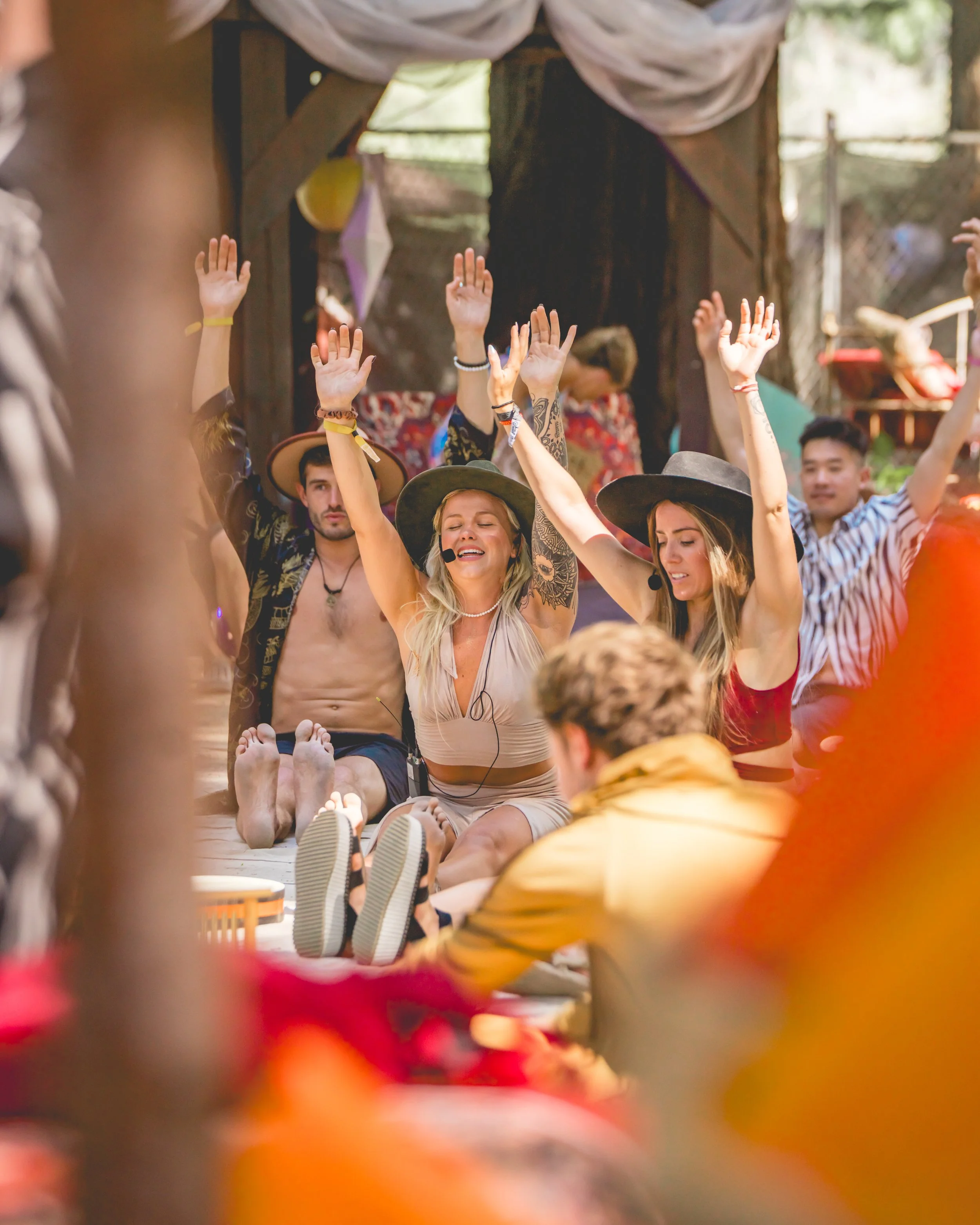 Group of young adults sitting on the ground, raising their hands in celebration, wearing summer clothes and hats, with a rustic outdoor setting in the background.