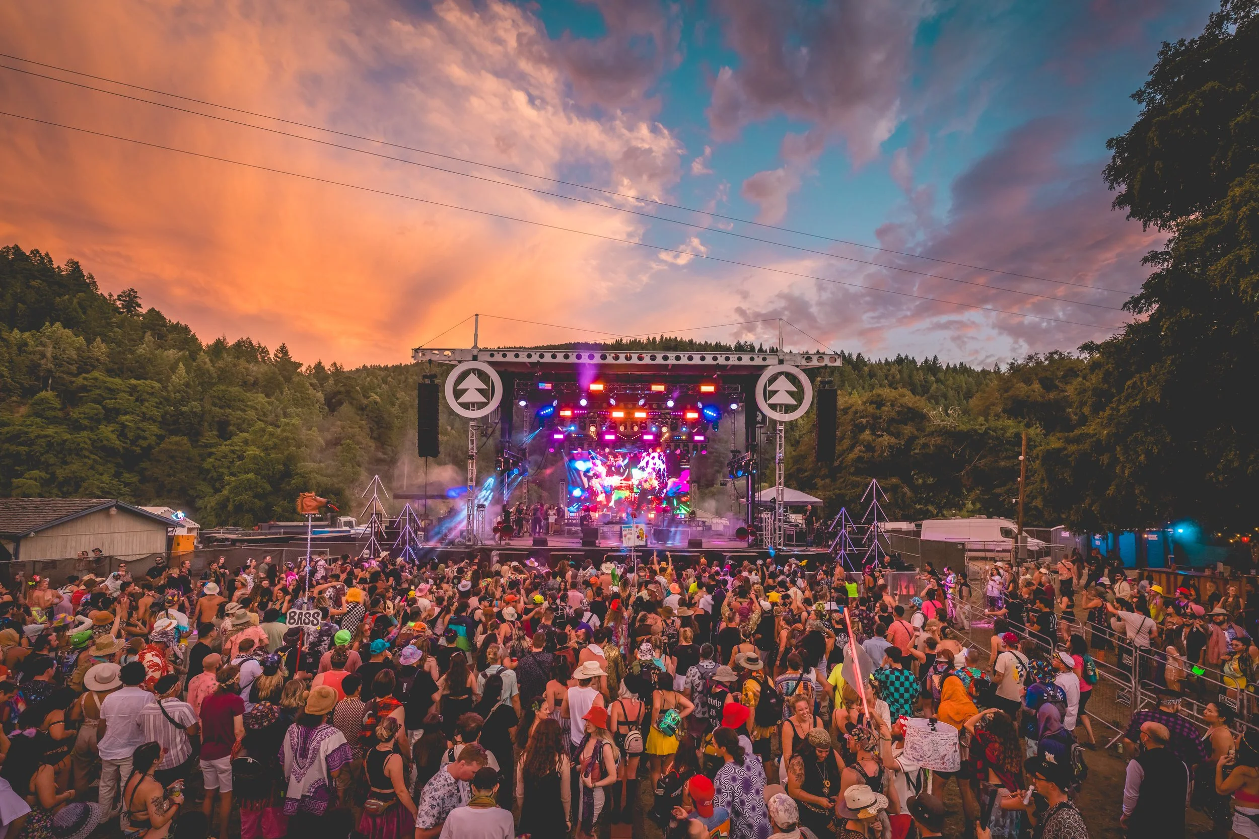 A vibrant outdoor concert at sunset with a large crowd watching a colorful stage surrounded by lush trees and a partly cloudy sky.