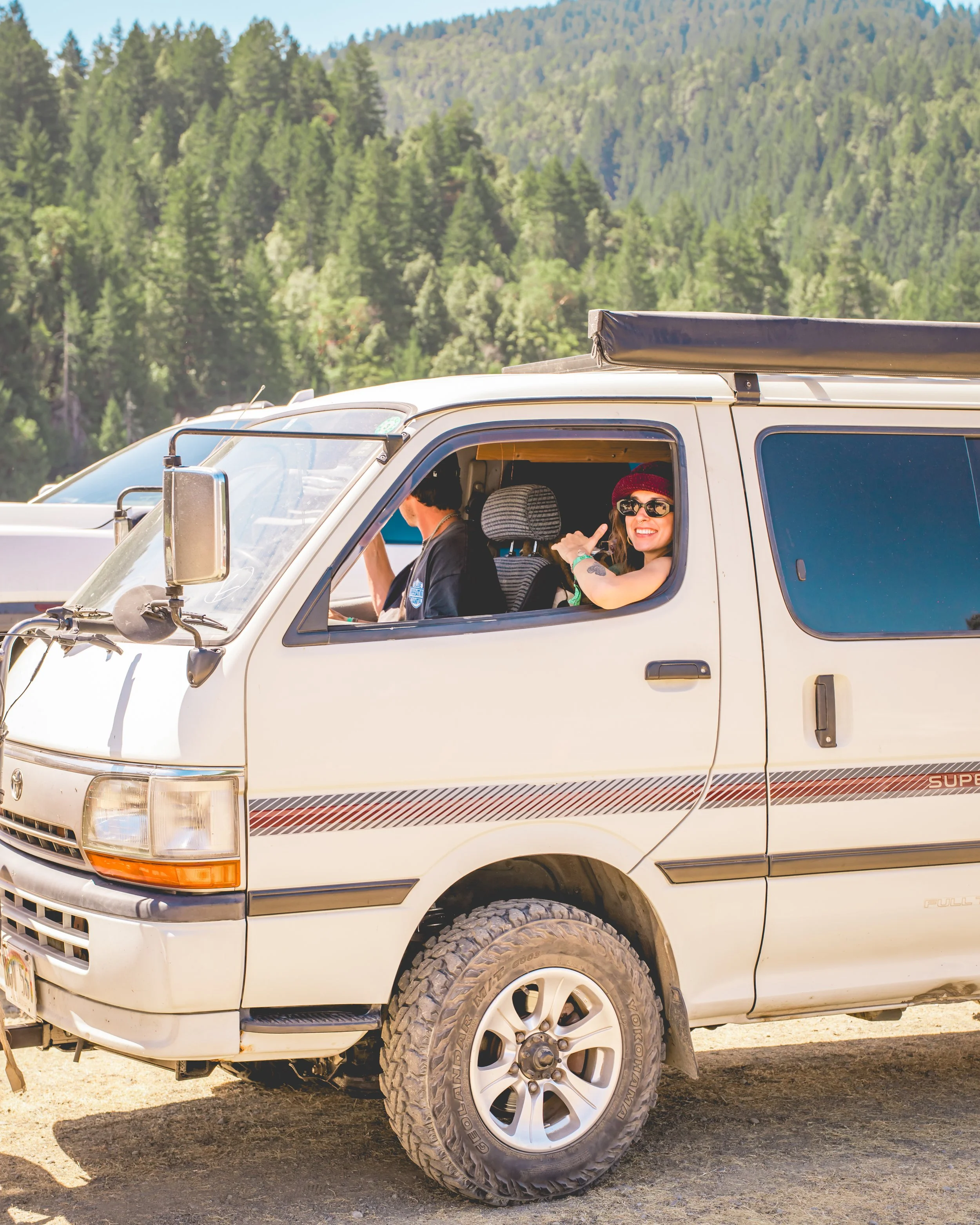 A smiling woman wearing sunglasses and a red beanie riding in the passenger seat of an off-road van in a mountainous area with pine trees.