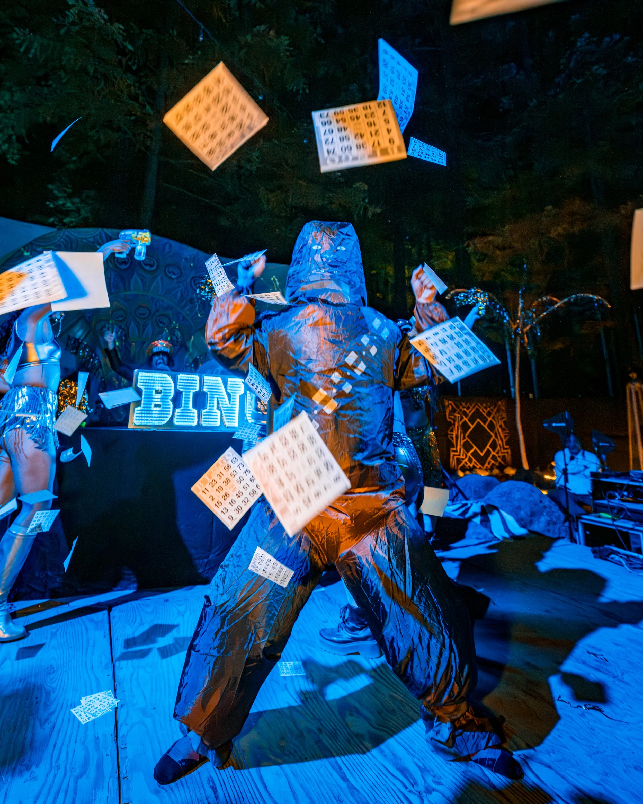 Person in a shiny, metallic outfit with a hood, standing on a stage with lottery tickets flying around during a bingo event at night.