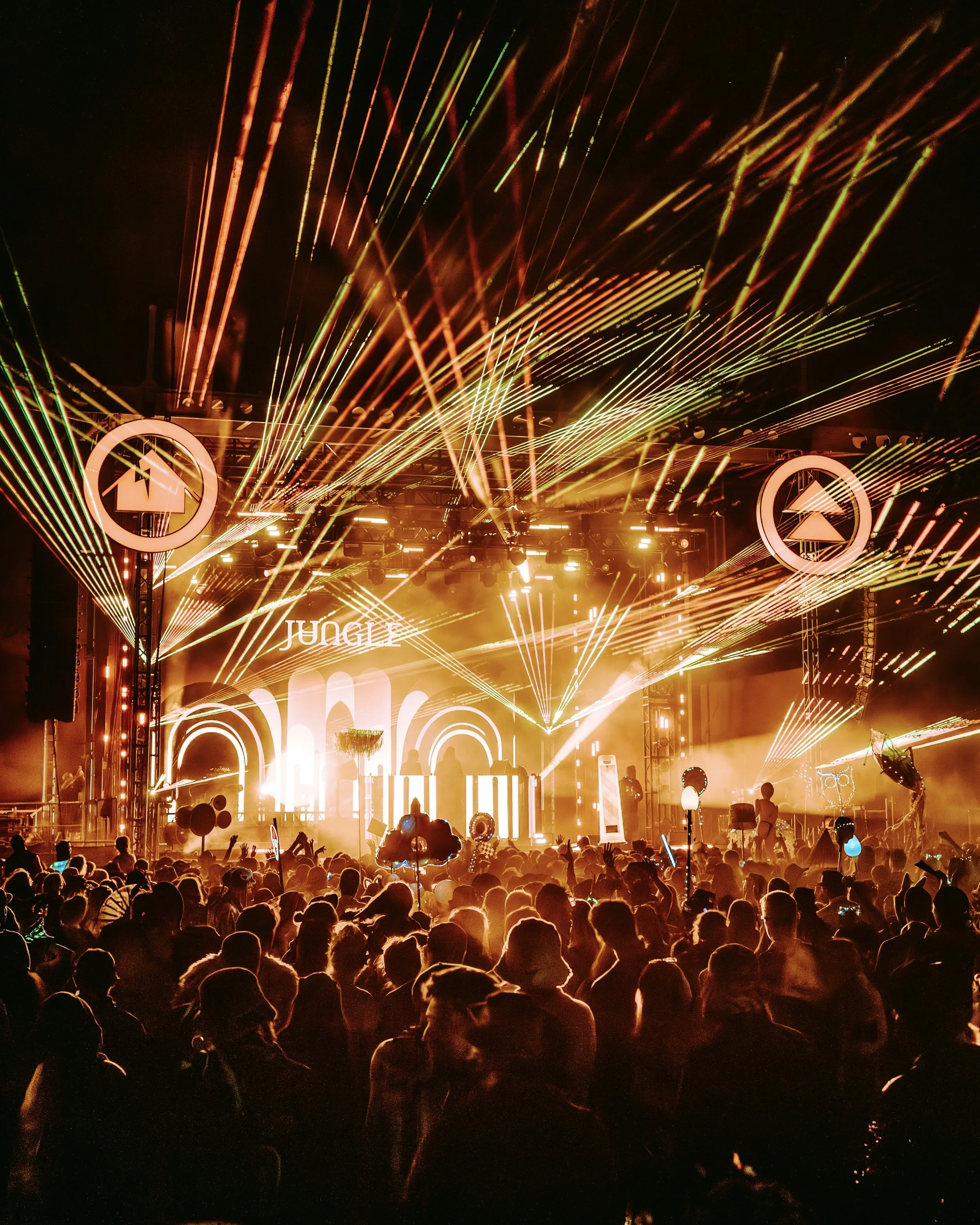 A lively concert scene at night with a large crowd, bright stage lights, laser beams, and DJ equipment on stage.