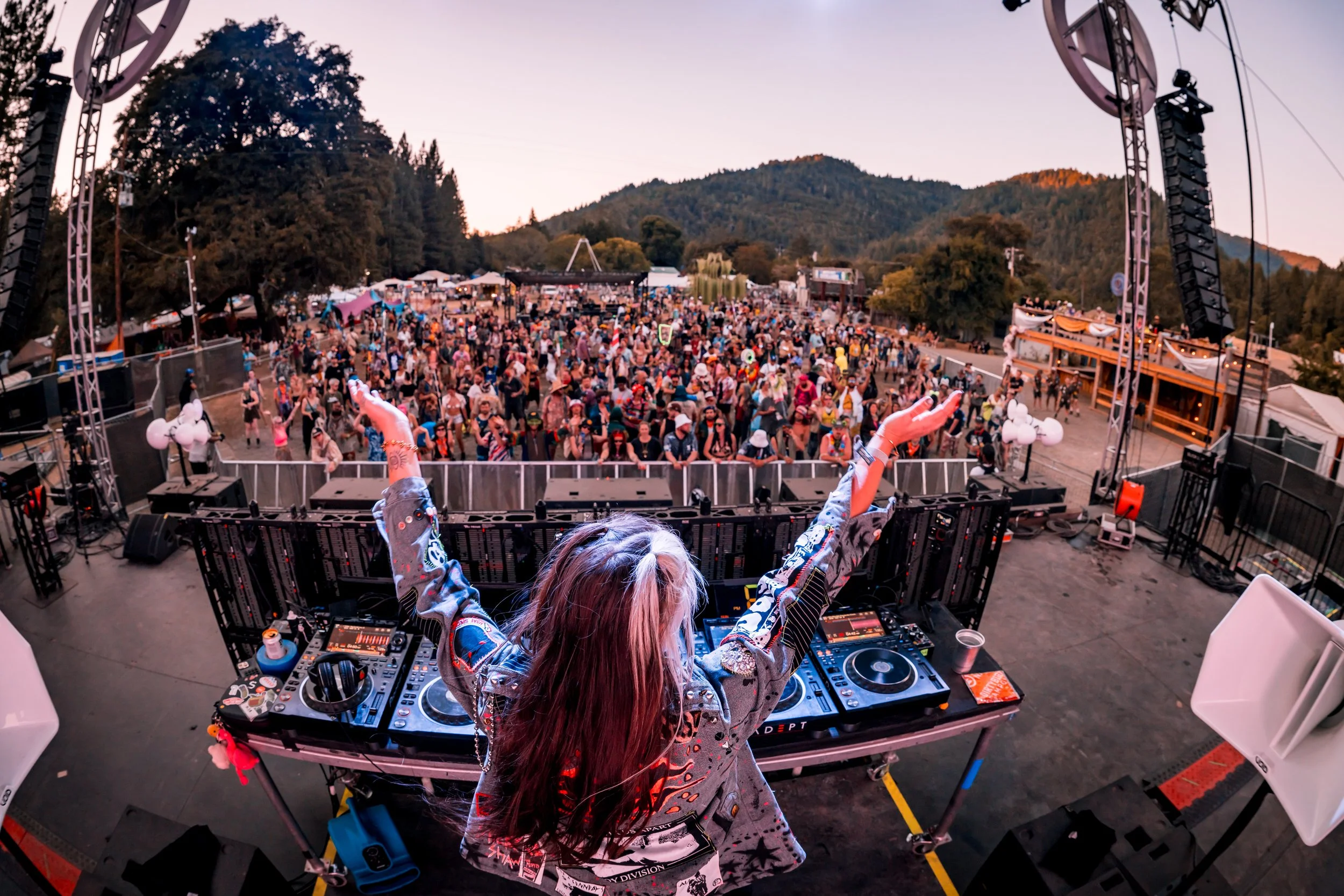 Female DJ with long hair and arms raised, performing on stage at an outdoor music festival with a large crowd and scenic mountains in the background.