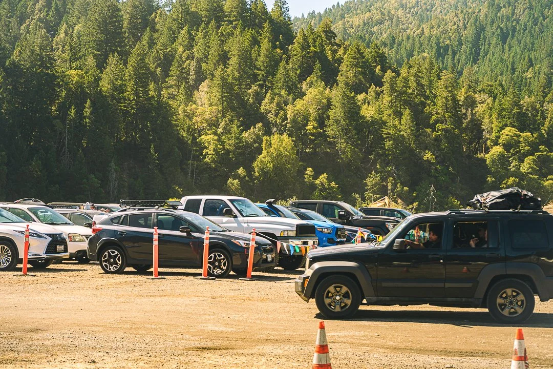 Reserved Parking: cars parked in a dirt lot marked with orange cones, with forested hills behind, at Northern Nights.
