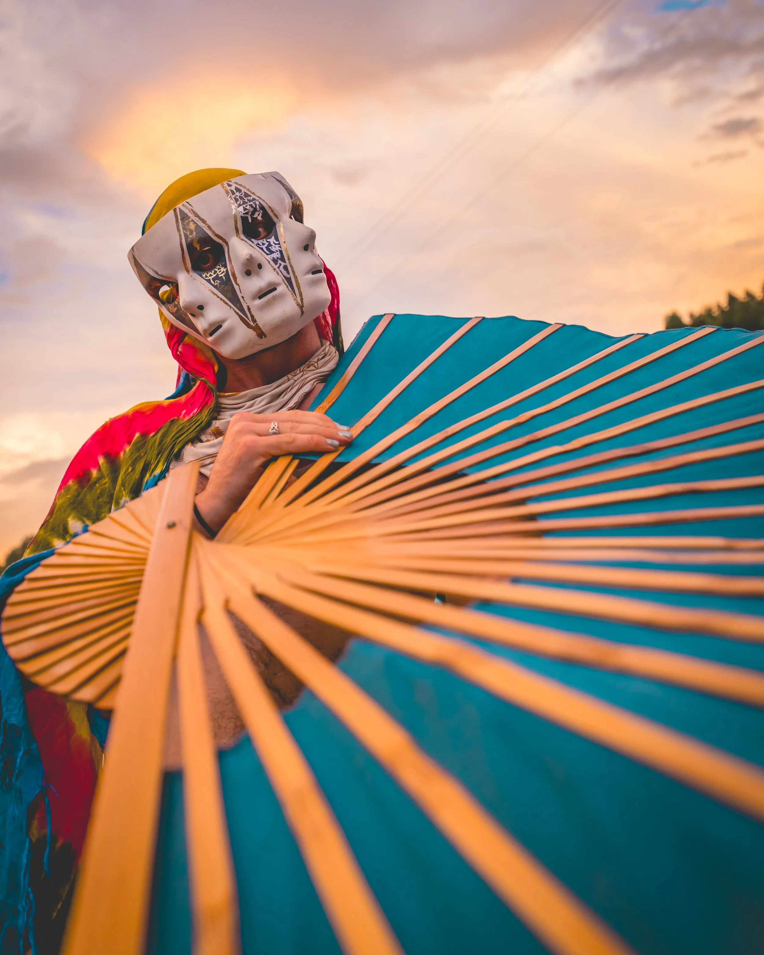 Person wearing a mysterious mask holding a blue and beige paper fan during sunset or sunrise.