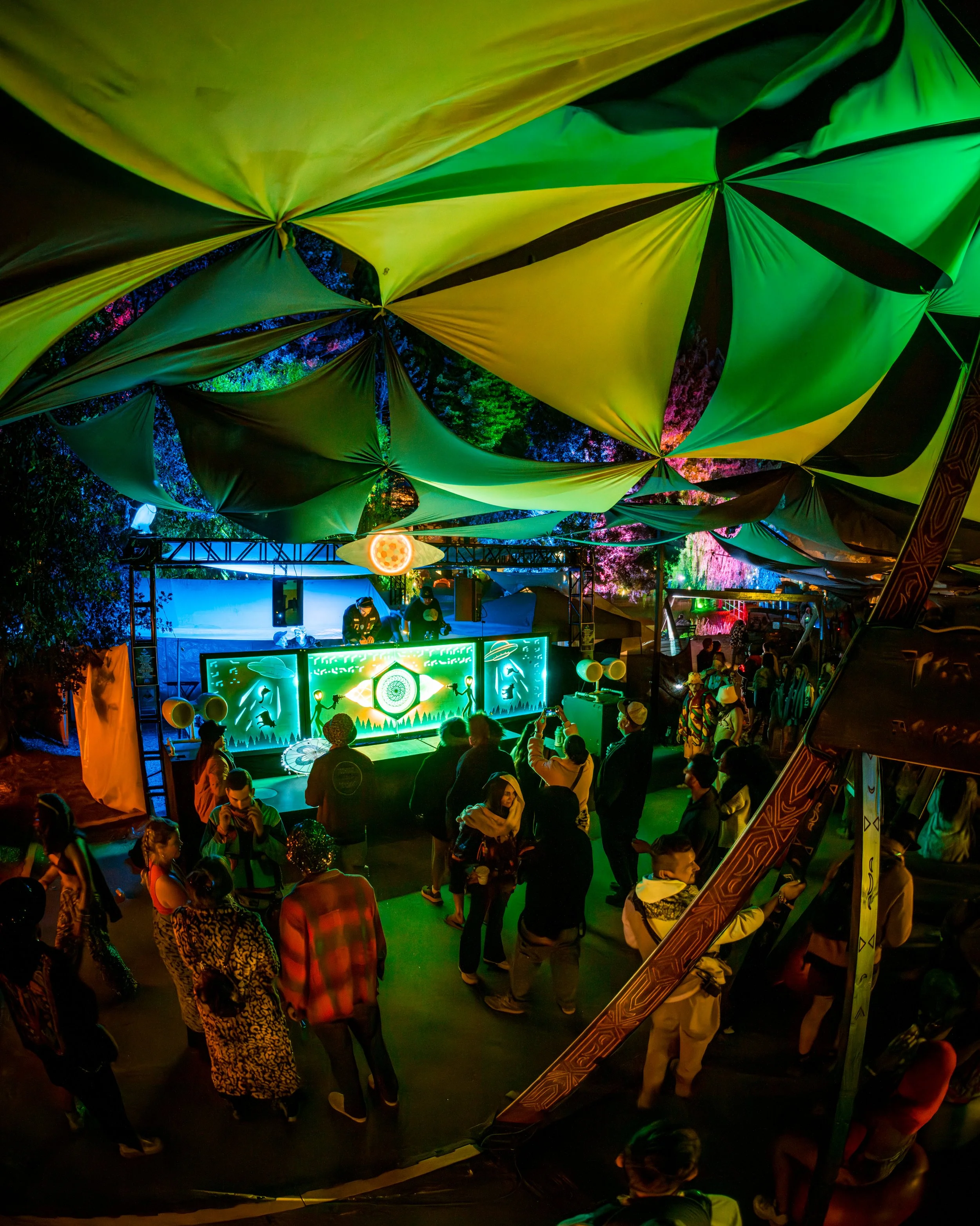 A vibrant outdoor night scene with a crowd of people dancing and enjoying a musical performance under a large, colorful fabric canopy with green, yellow, and black sections, lit by neon and stage lights.