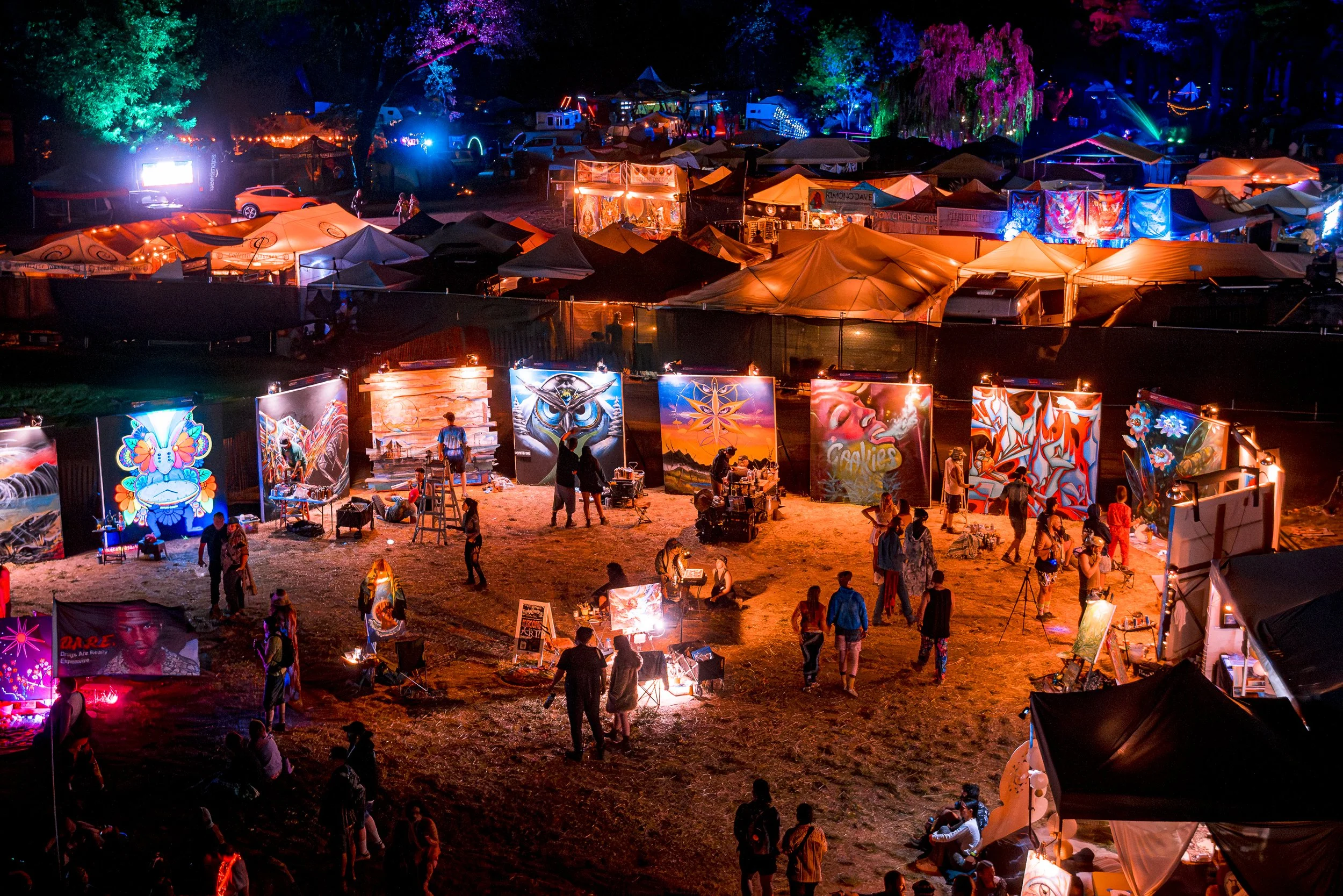Night scene of an outdoor art festival or fair with illuminated artworks and tents, people walking and viewing art.