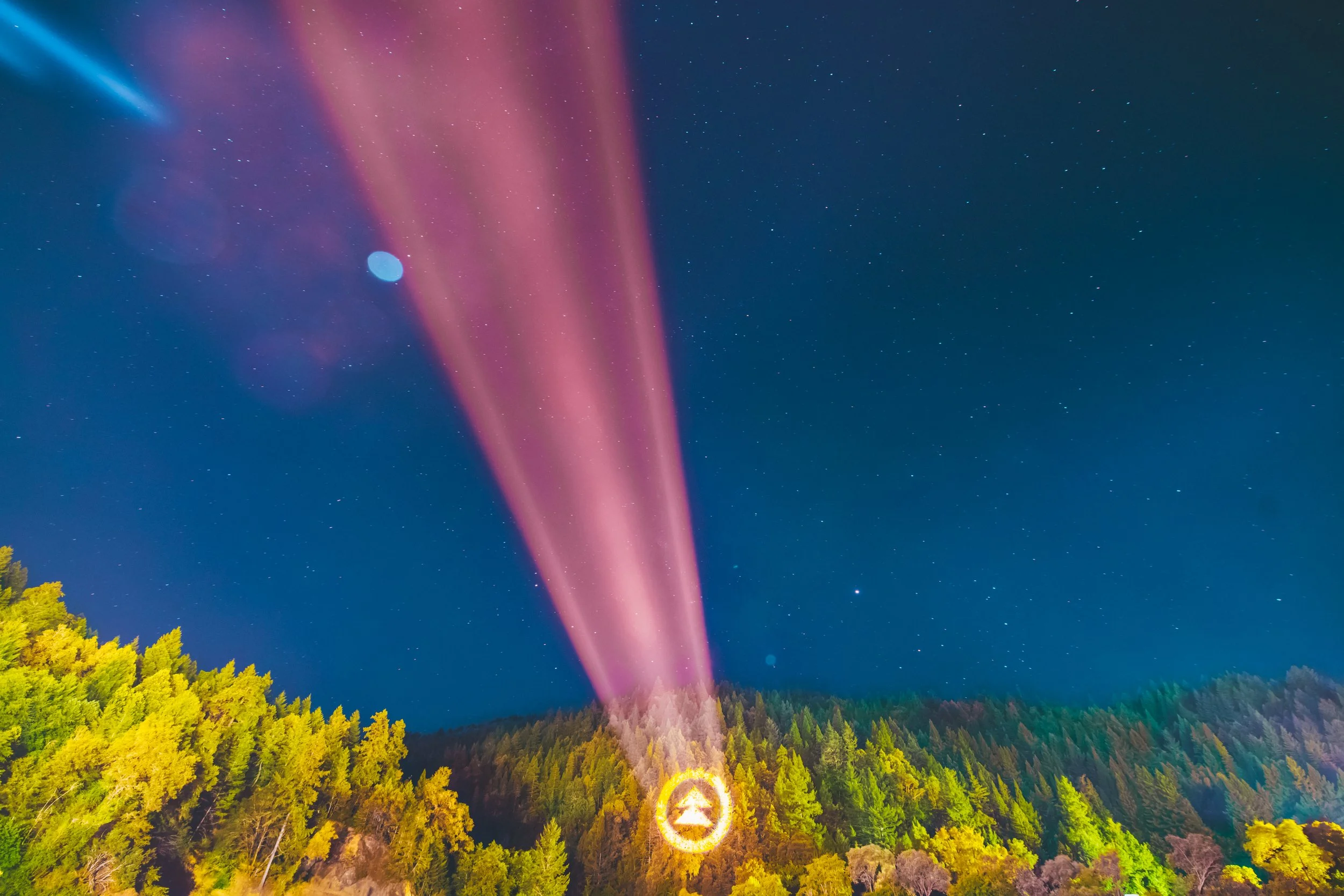 Night sky with stars and colorful light beams over a forest, with a bright circular logo at the base of the trees.