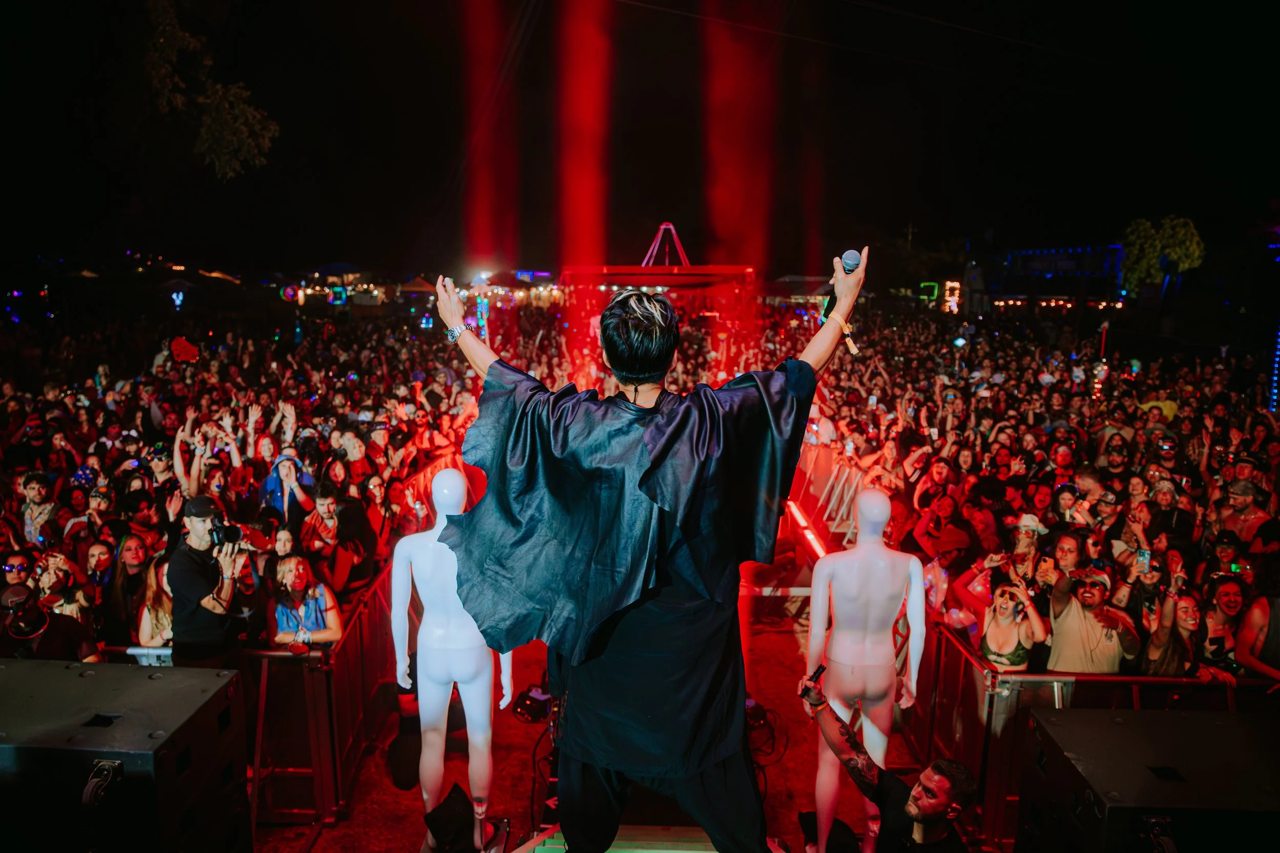 Performer on stage with arms raised at a large outdoor music festival at night, facing a crowd of thousands of concertgoers.