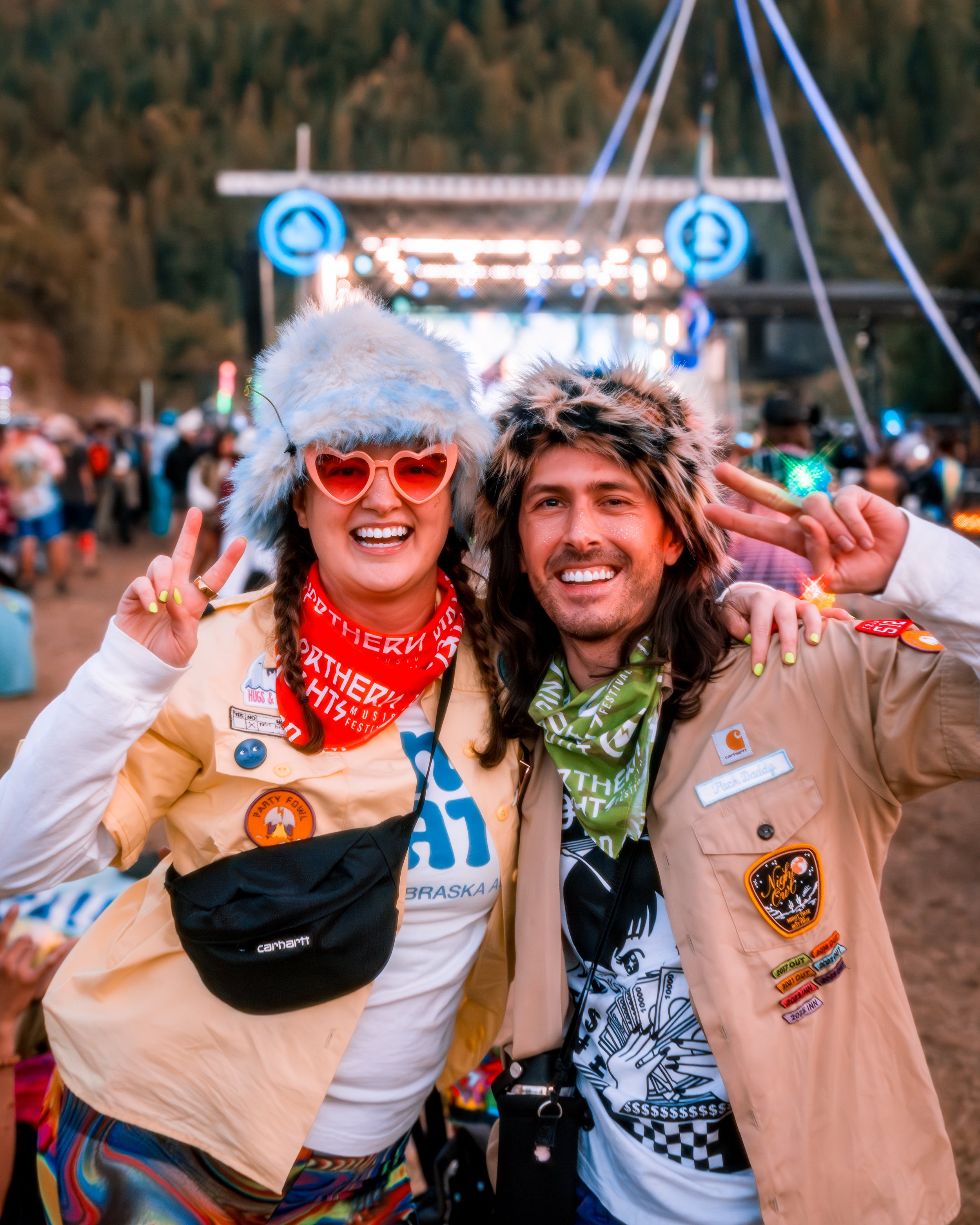 Two people smiling at a music festival, wearing fur hats and colorful outfits. The person on the left is making a peace sign, wearing heart-shaped sunglasses, and has a crossbody bag. The person on the right is also making a peace sign and has a came